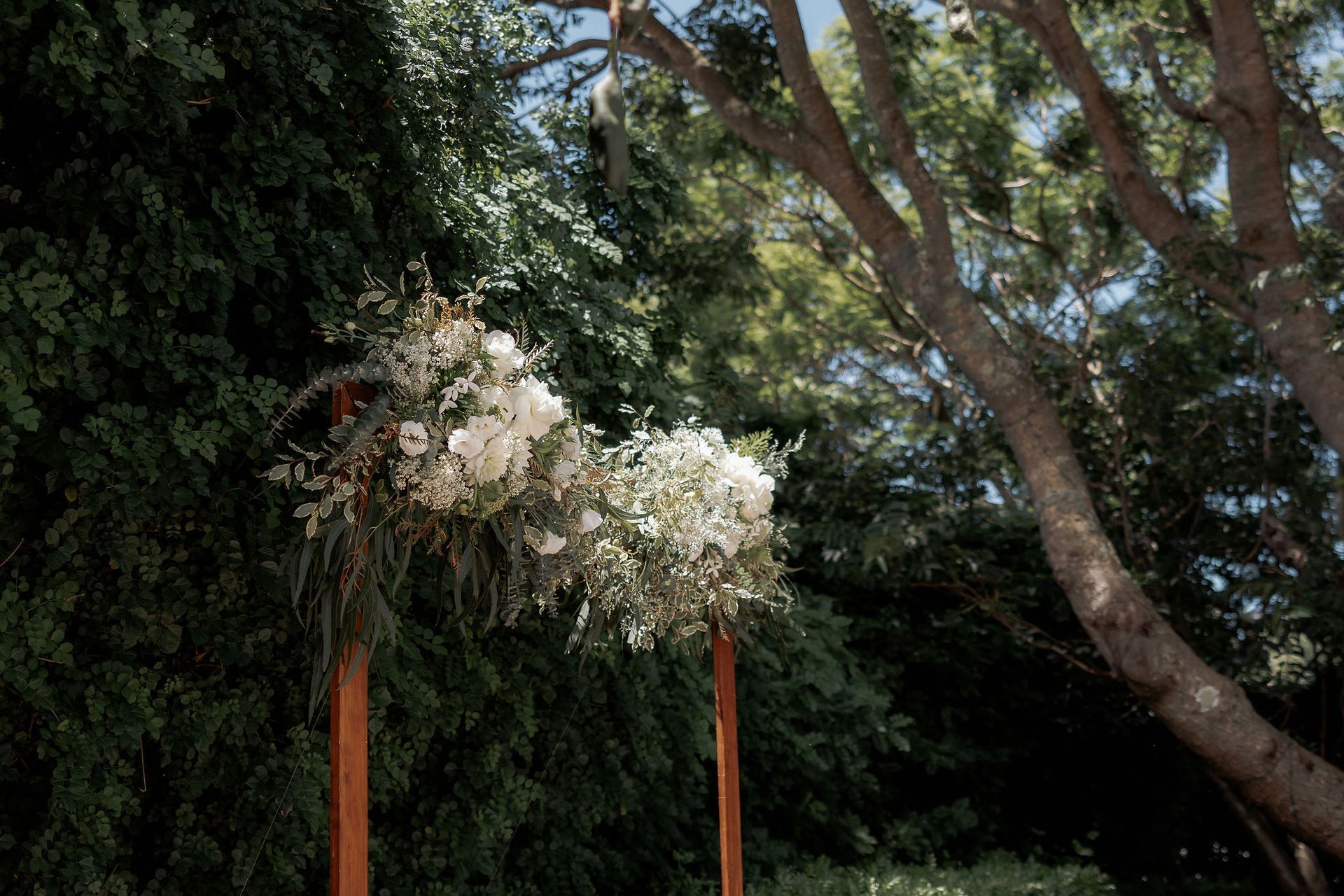 Floral arrangements on wooden posts, set outdoors against greenery and a tree.