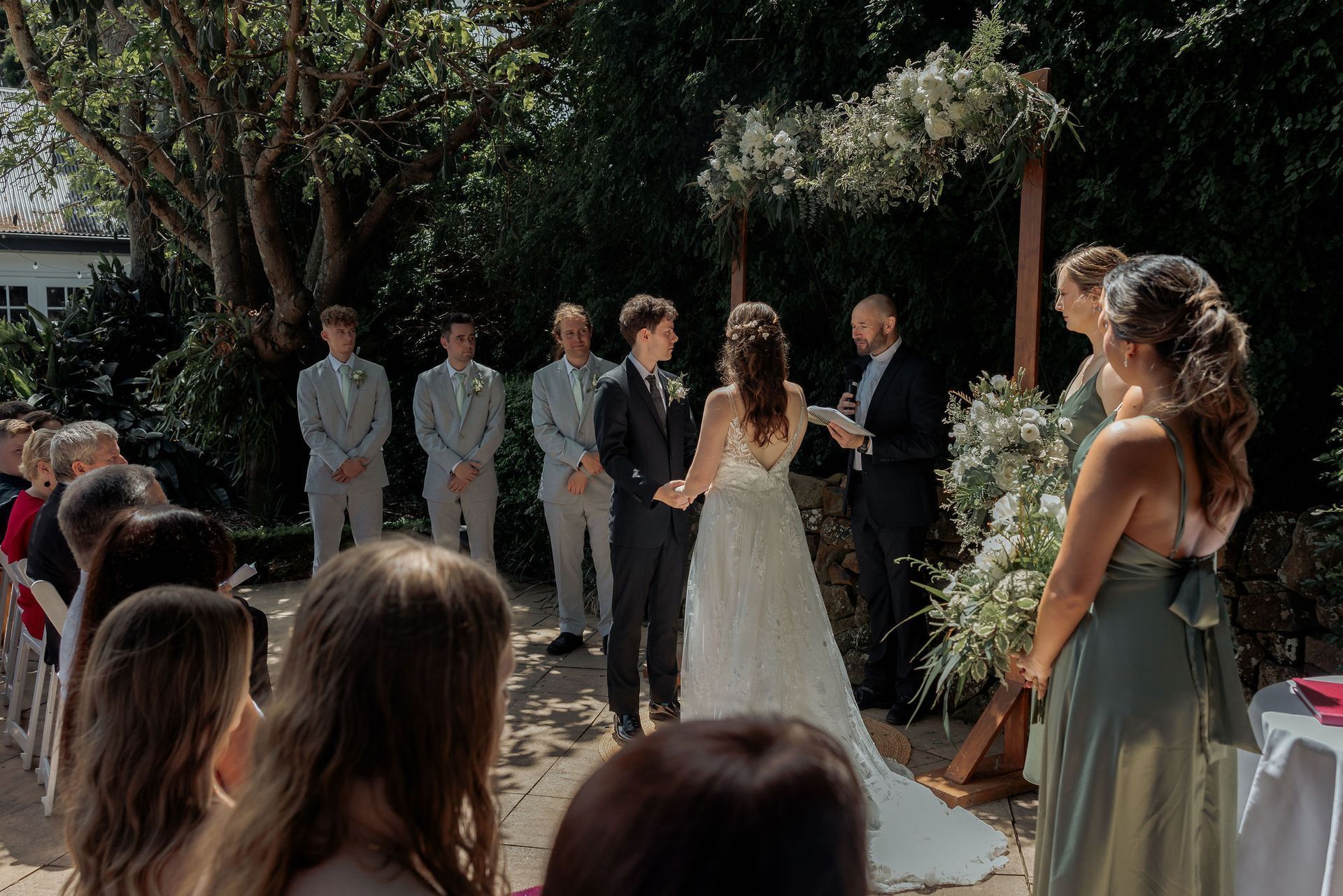 Wedding ceremony: Couple exchanging vows outdoors, officiant, wedding party, guests. Green foliage, sunny day.