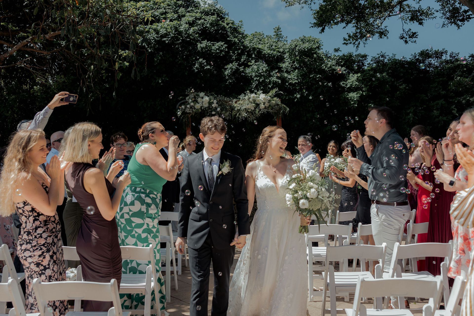 Newlyweds walk aisle, showered with bubbles, guests clapping, sunny outdoor wedding.