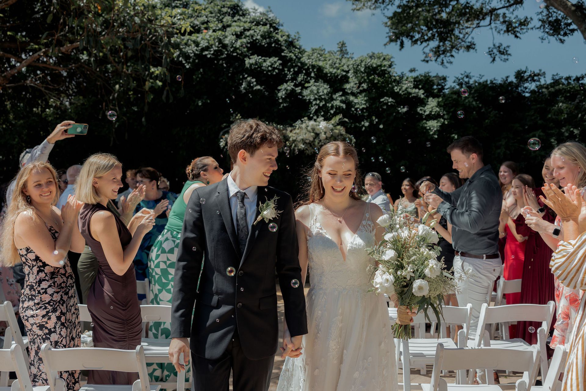Newlyweds walk down aisle lined with applauding guests. Sunny outdoor wedding.