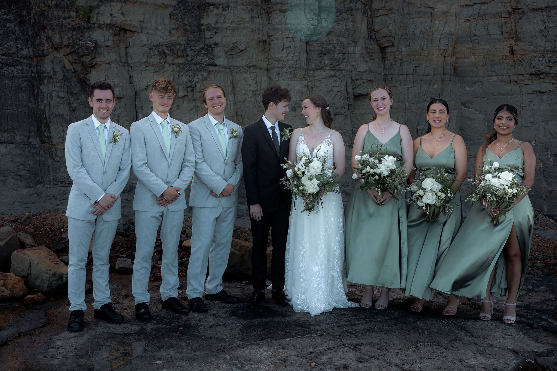 Wedding party: Bride and groom in front, bridesmaids in green dresses, groomsmen in light suits, all near a rocky wall.