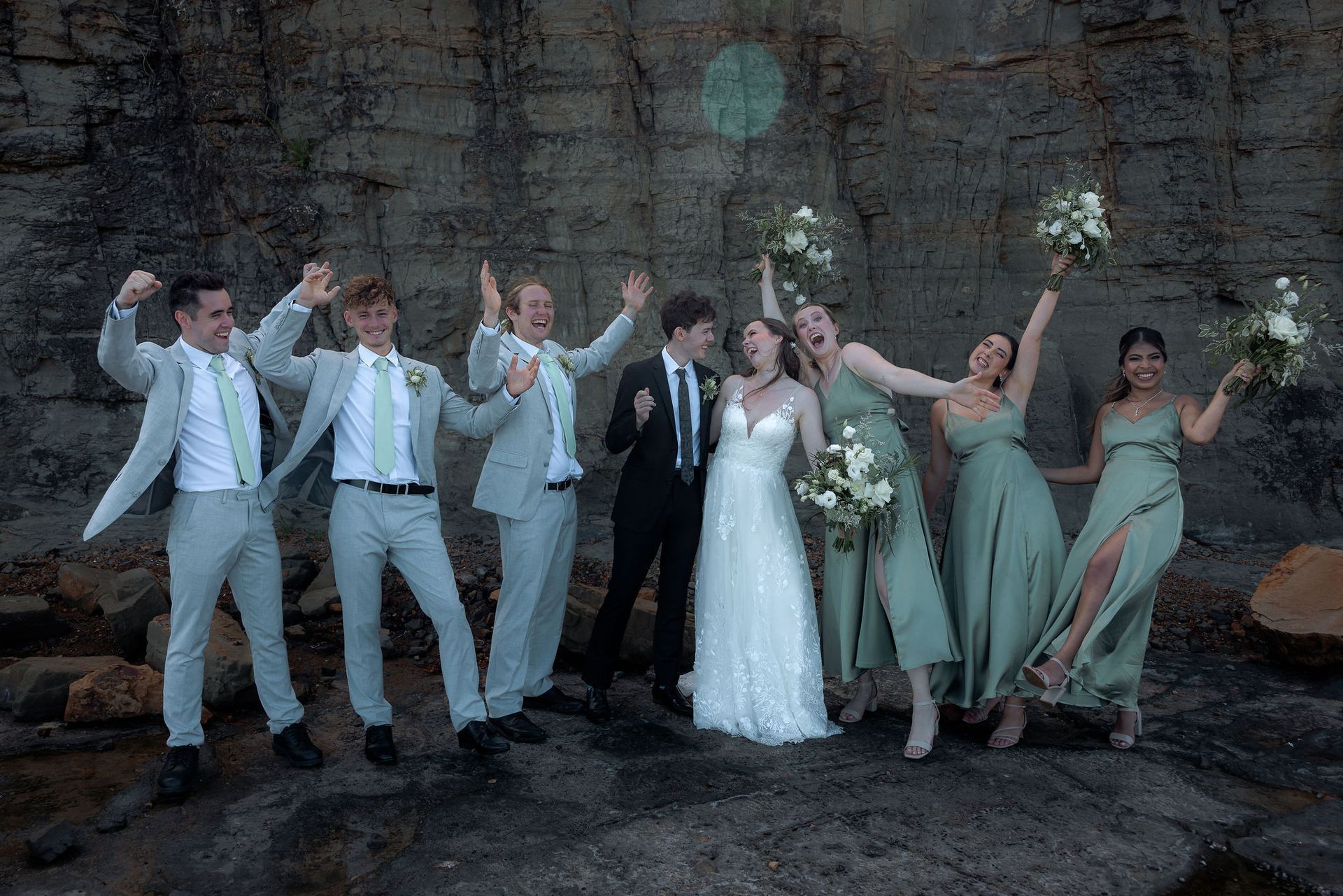 Wedding party poses, all celebrating. Bride and groom in center, surrounded by wedding party, against rocky cliff.