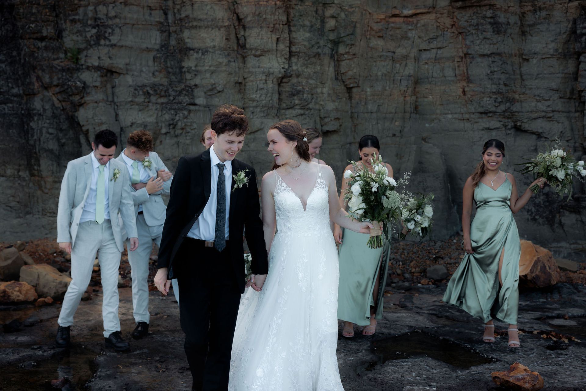 Wedding party: Bride and groom smiling, walking, followed by bridesmaids and groomsmen near a rock cliff.
