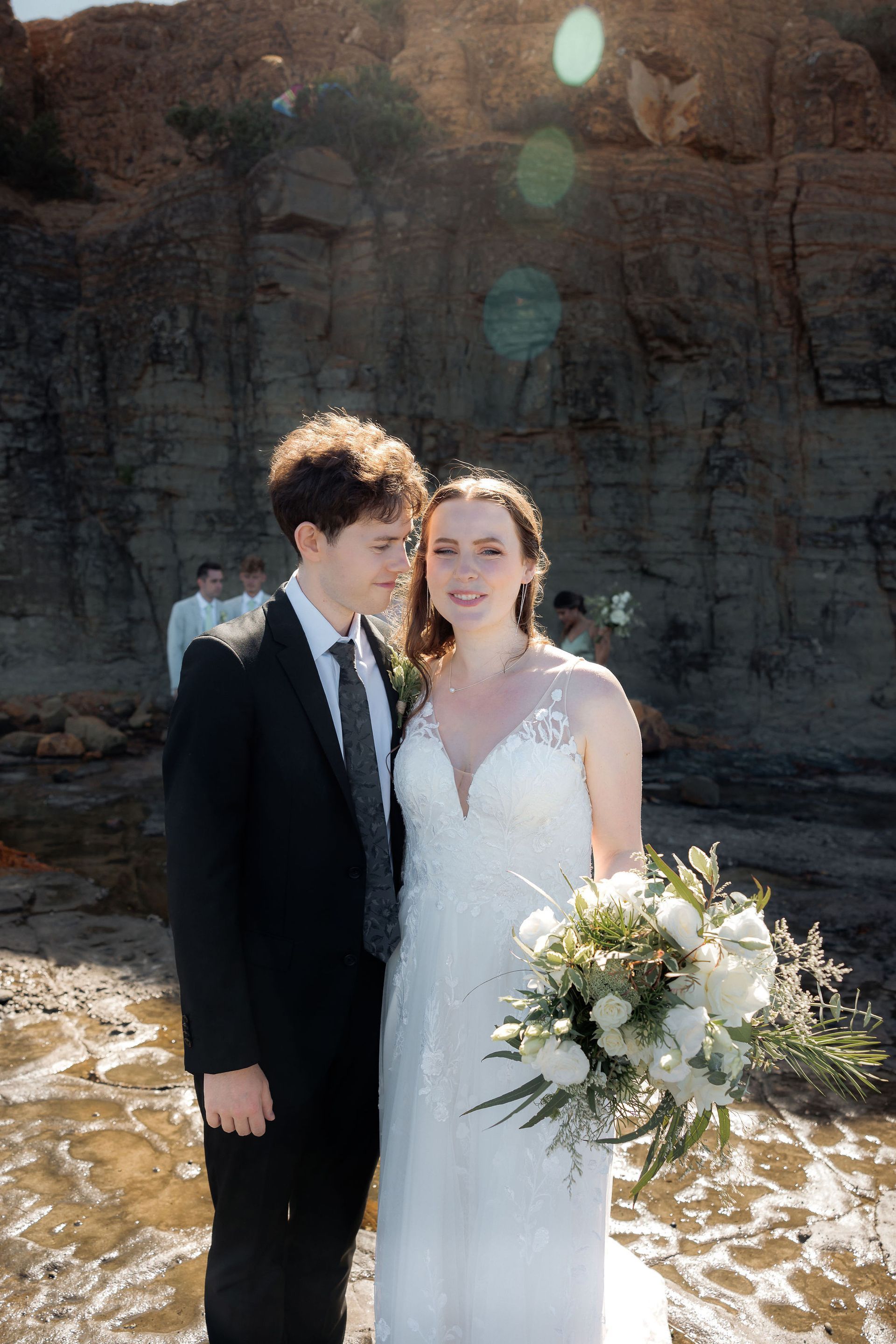 Wedding couple poses on rocky shore; man in black suit looks at woman in white dress holding flowers.