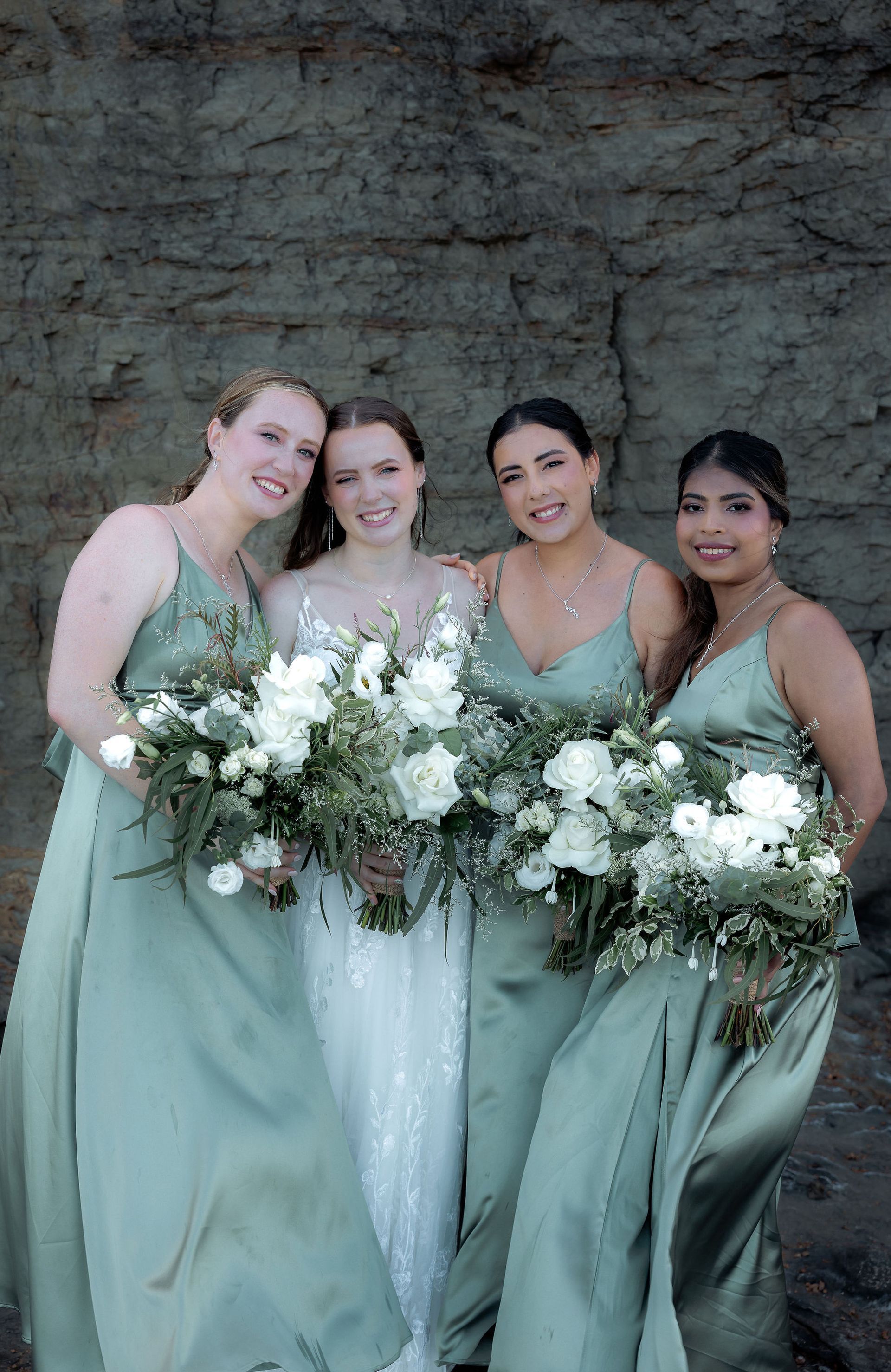 Bride and bridesmaids in sage green dresses, smiling, holding white bouquets, against a rock wall.