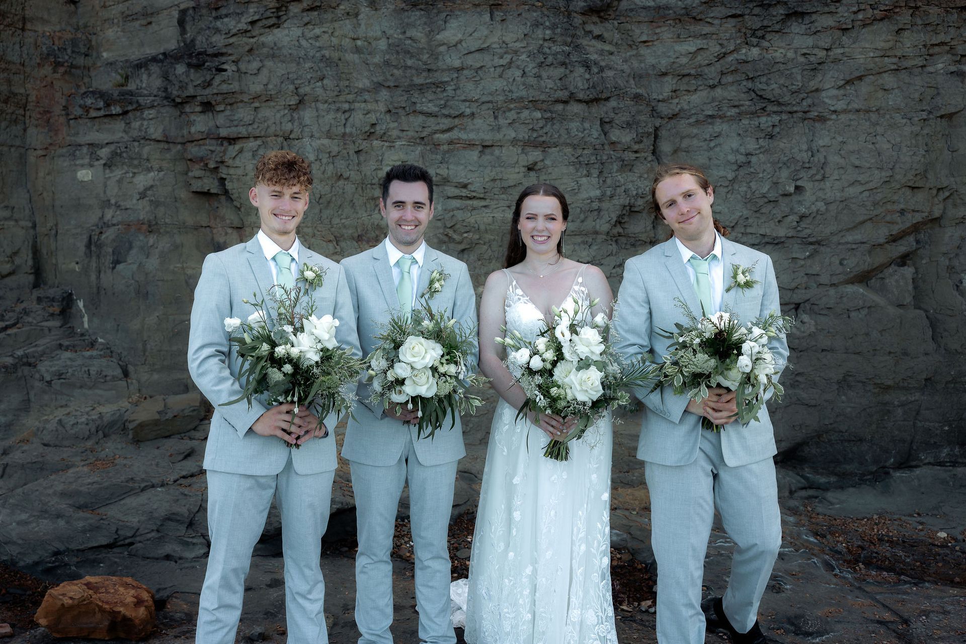 Bride and groom with two groomsmen holding bouquets, smiling in front of a rock wall. Light suits, dresses.