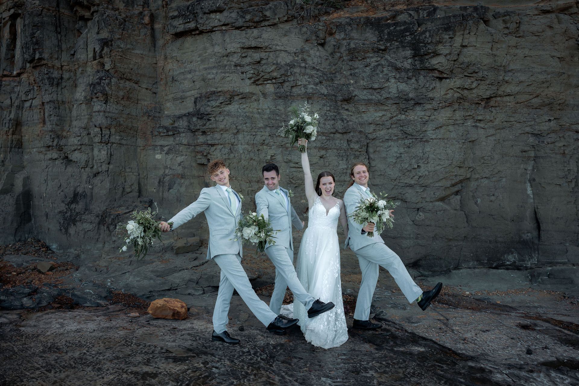 Wedding party poses joyfully in front of a rock face. Bride in white, groomsmen in light suits, holding bouquets.