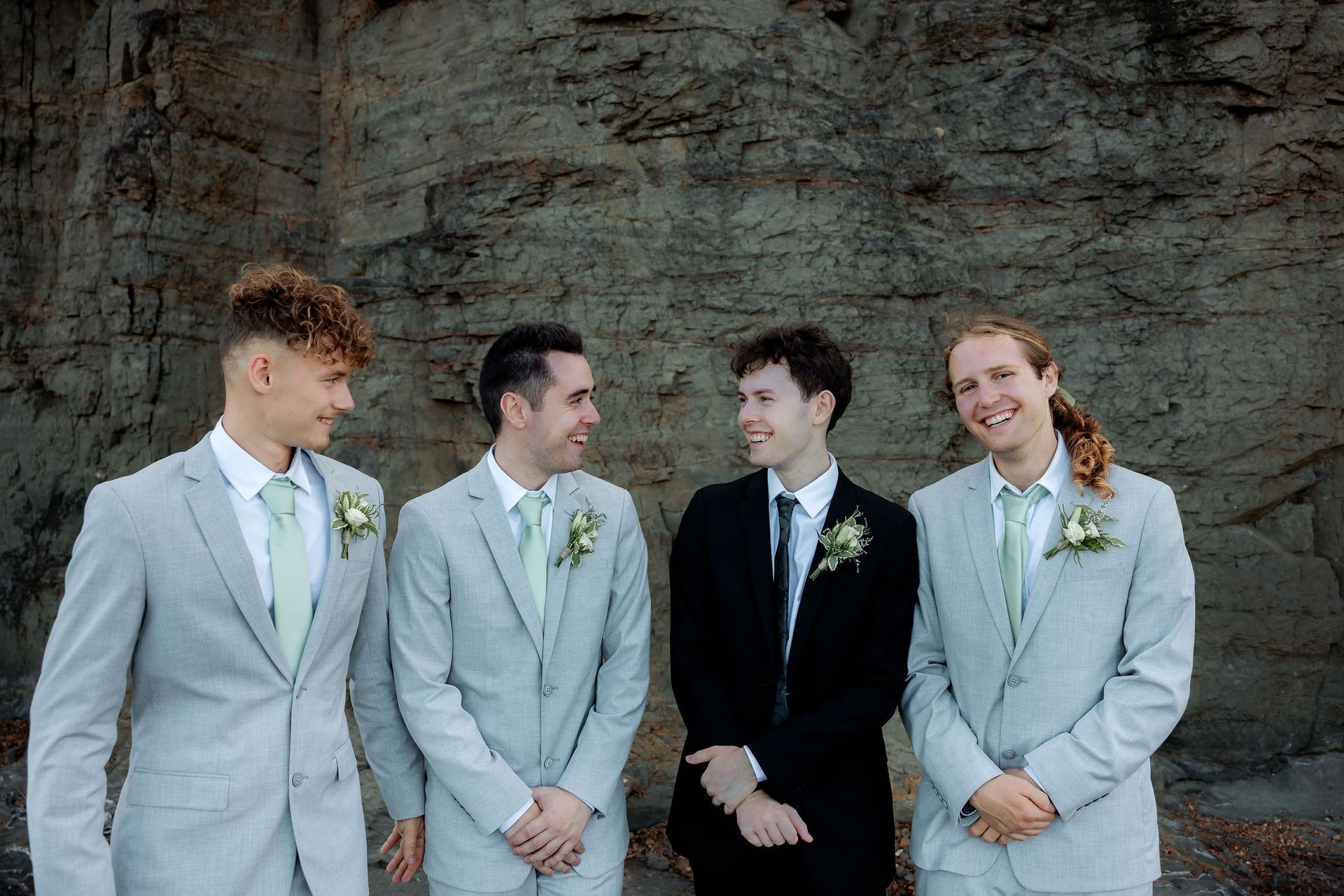 Four groomsmen pose in front of a textured stone wall. Three in gray suits and one in black, smiling at each other.