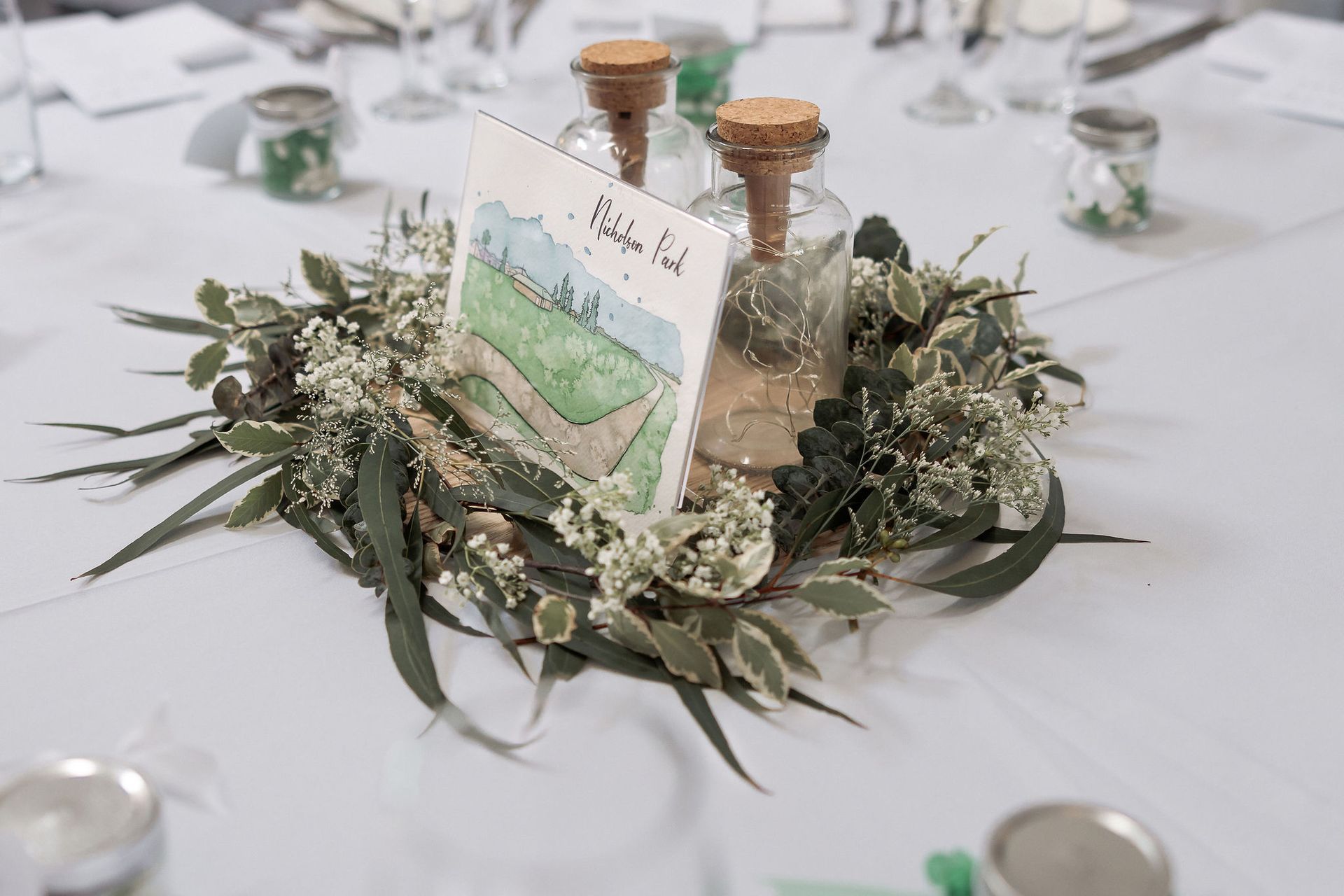Centerpiece with greenery, cork bottles, and a watercolor table card on a white tablecloth at a wedding.