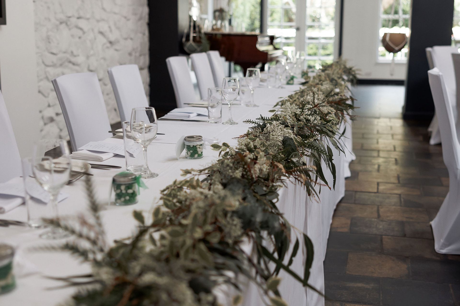 Long, white table set for a wedding, decorated with greenery, glasses, and white-covered chairs.