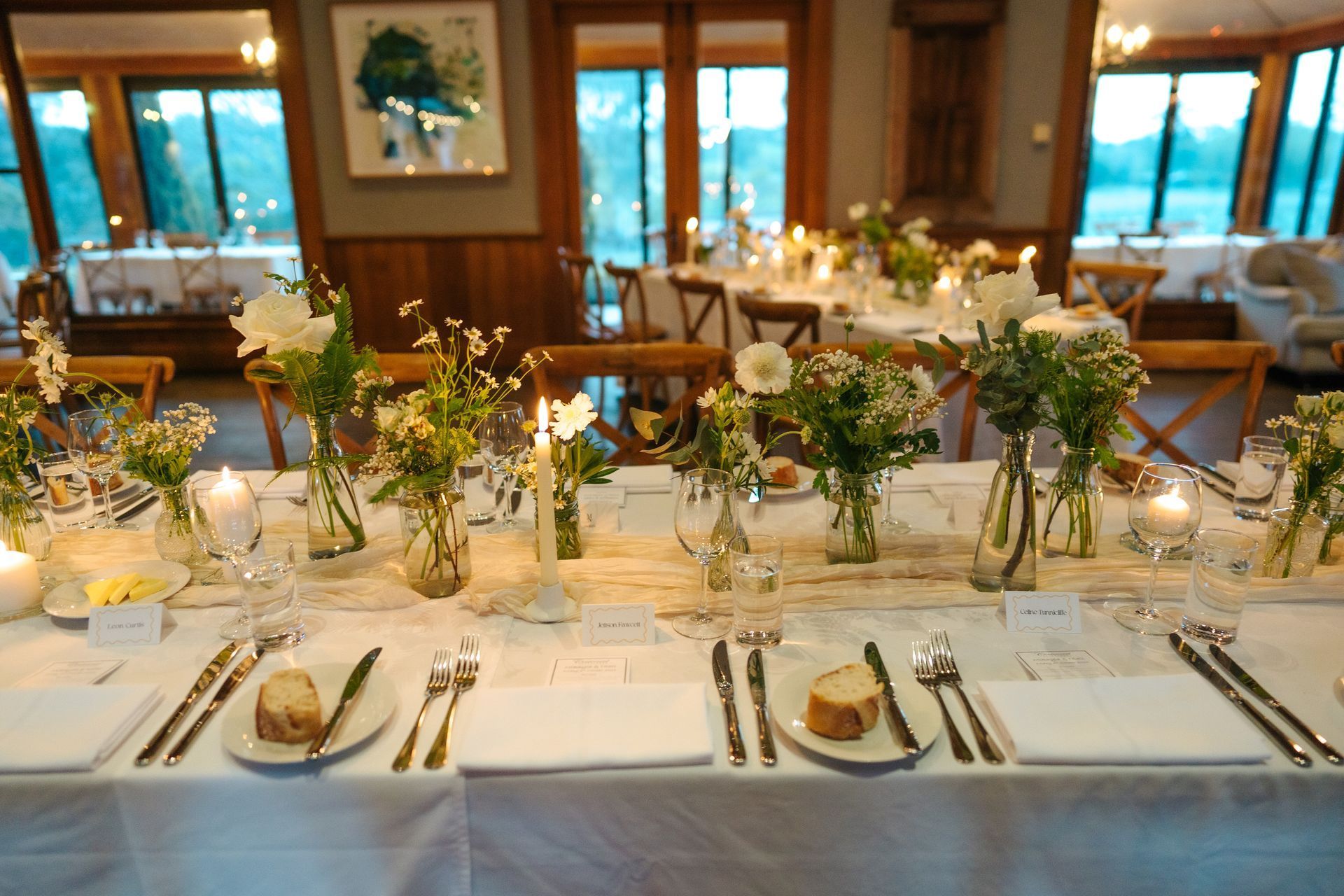 Formal dining table set for a celebration; white linens, flowers, candles, in a room with large windows.
