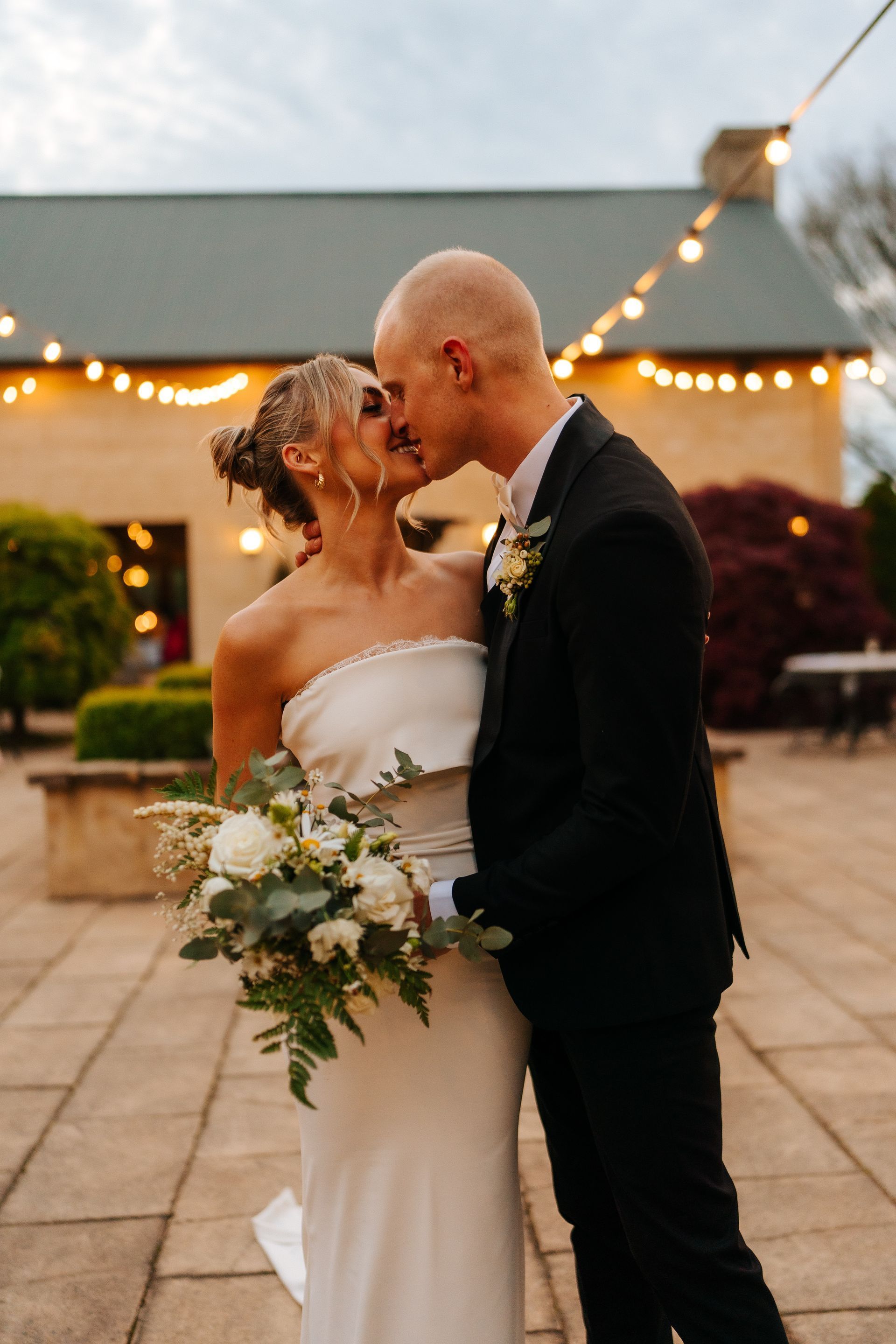Bride and groom kissing, outdoor wedding, woman in white gown with bouquet, man in black suit, string lights.