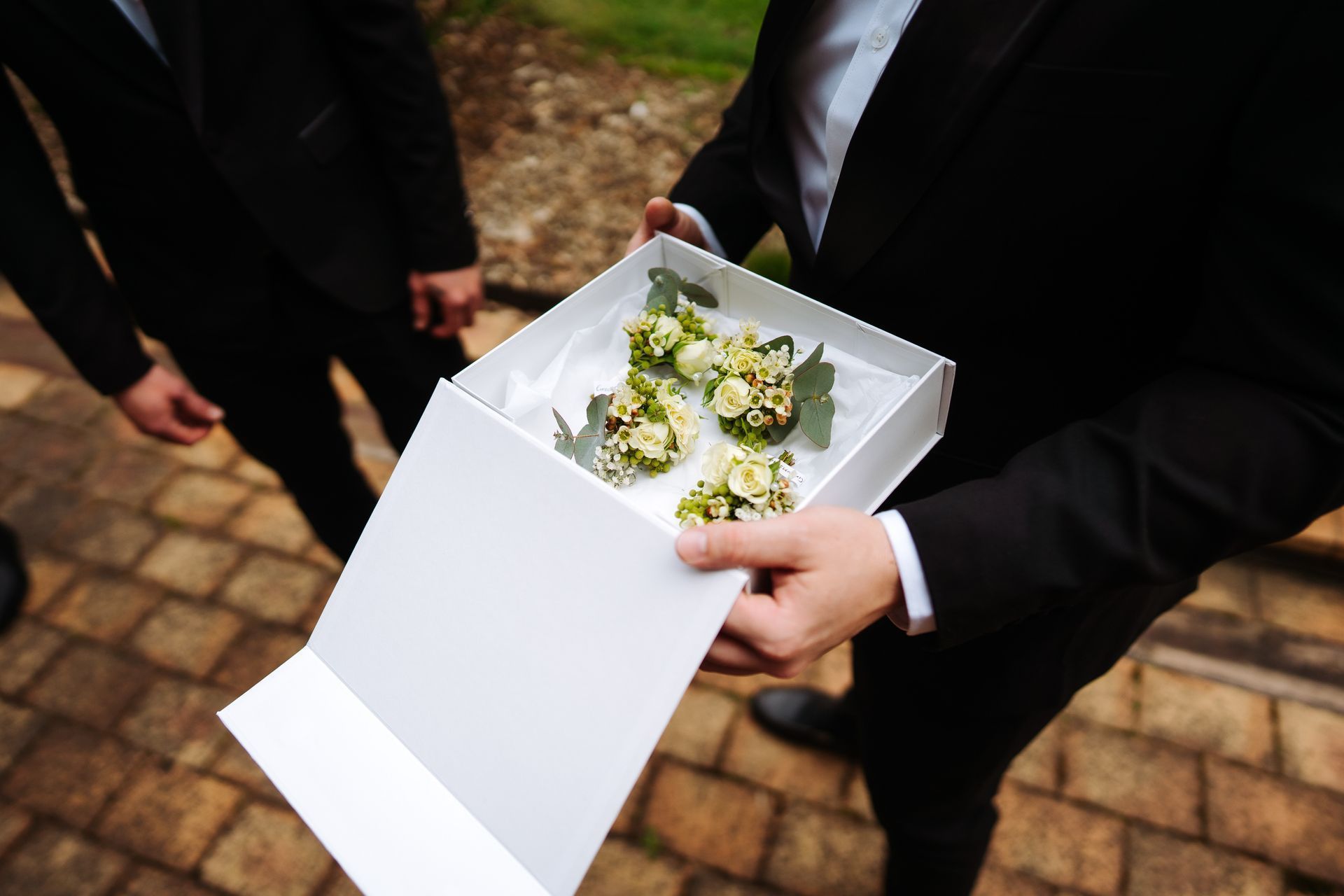 Man in suit holding a box of boutonnieres. Other men in suits stand nearby on a brick path.