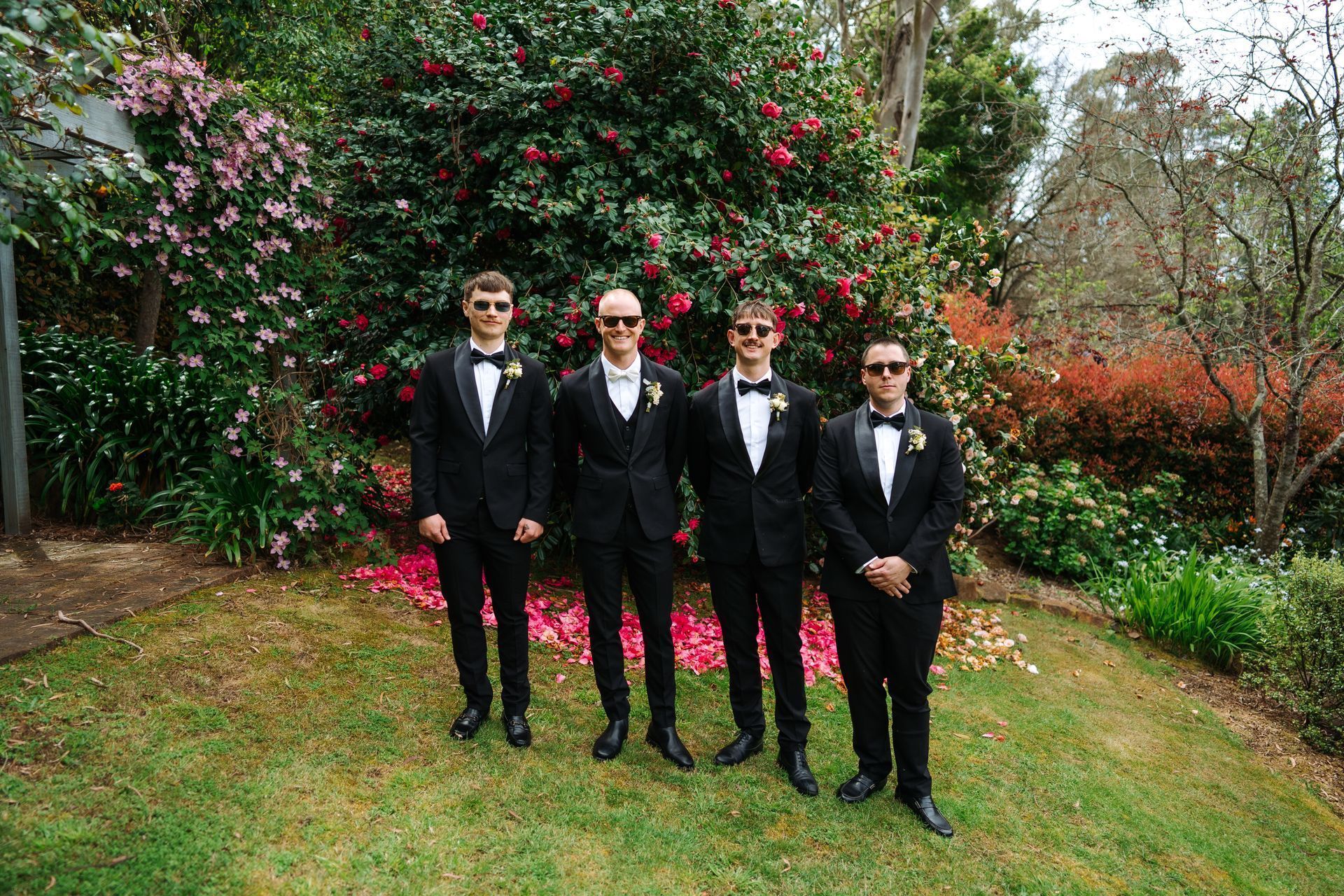 Four men in black tuxedos stand outside in front of a lush green bush and flowers.