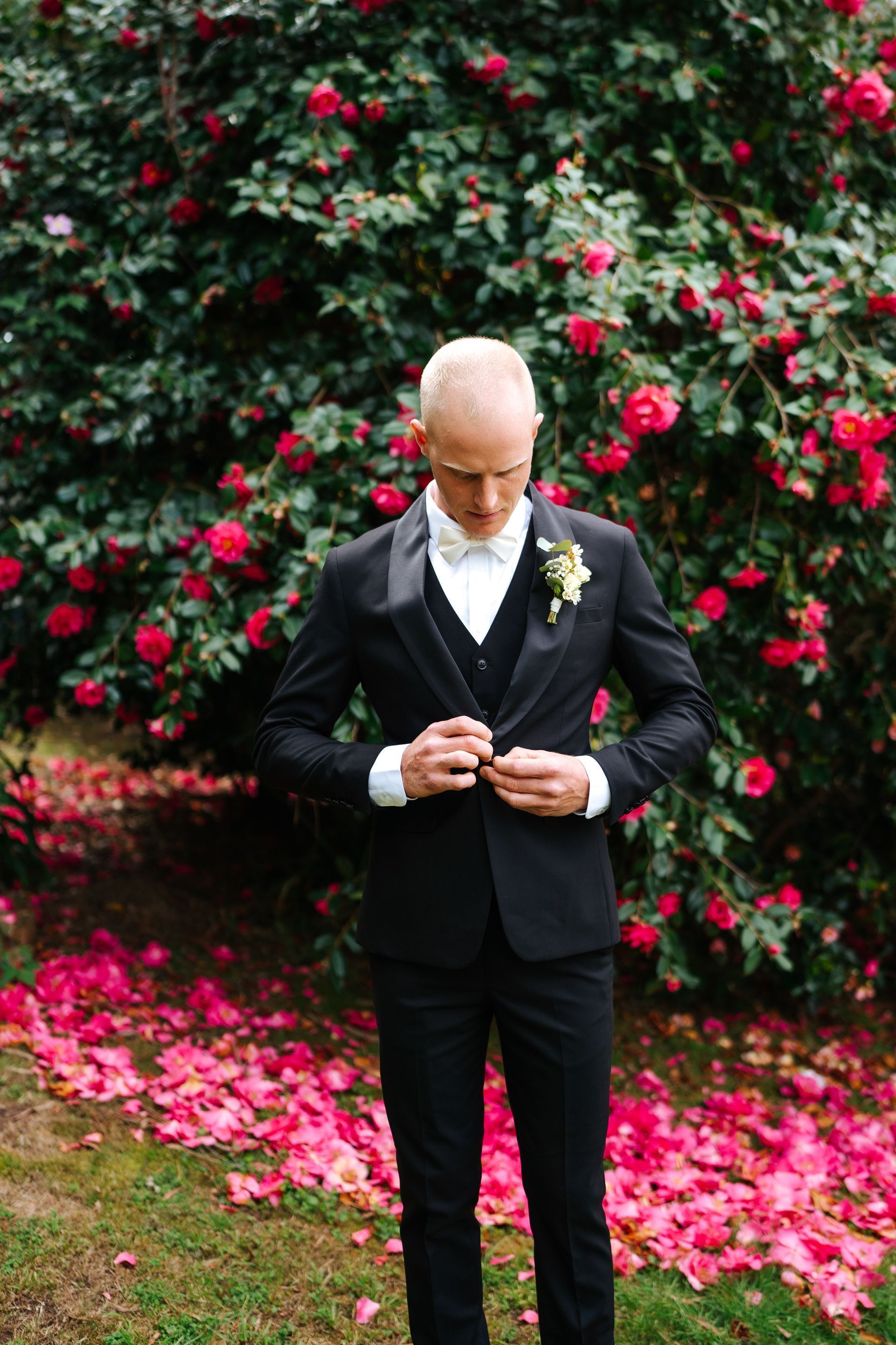 Groom in black tuxedo adjusting his jacket, standing in front of a bush with red flowers and fallen petals.
