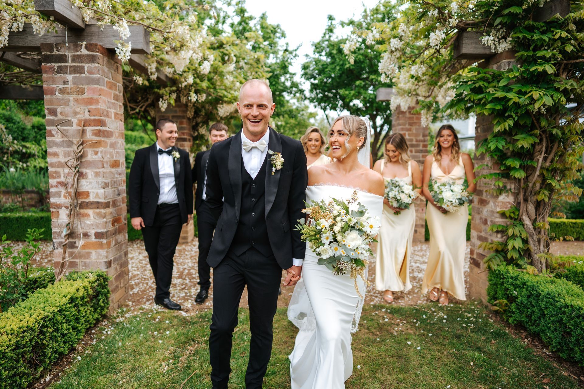 Newlyweds and wedding party walk down a garden path, smiling. Bride in white gown; groom in black tux.
