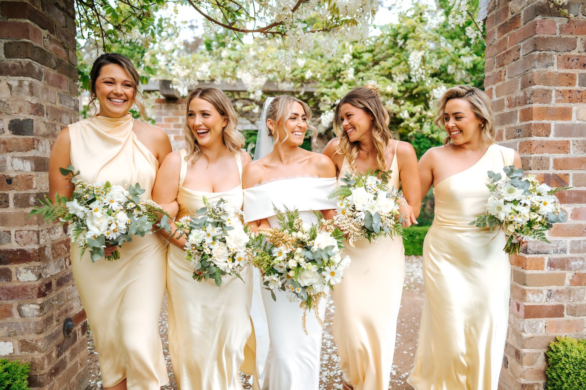 Bride with bridesmaids smiling, holding bouquets, wearing ivory dresses. Outdoor setting with brick archway.