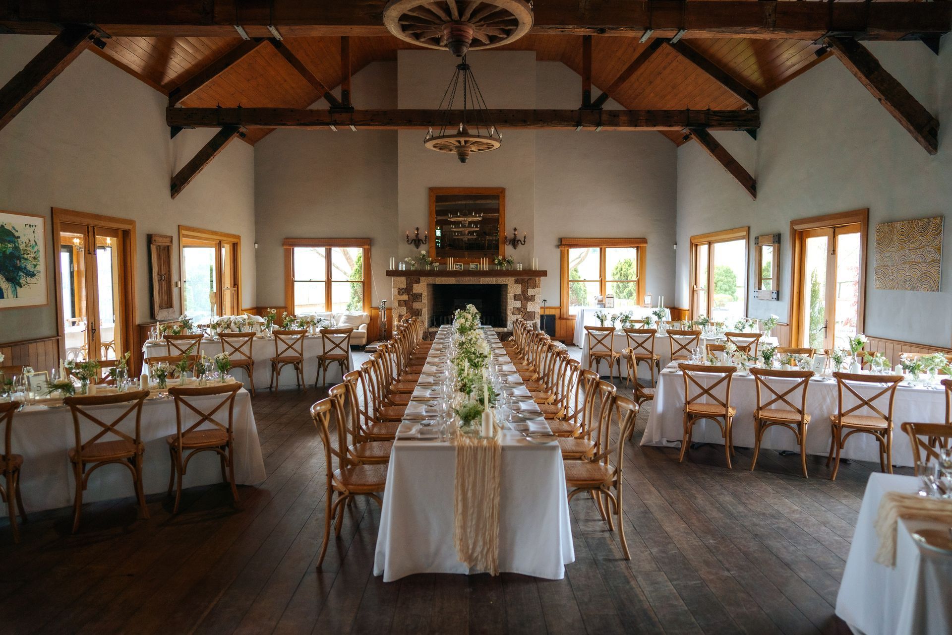 Wedding reception: Long table with white linens, wooden chairs, fireplace, and windows in a rustic room.