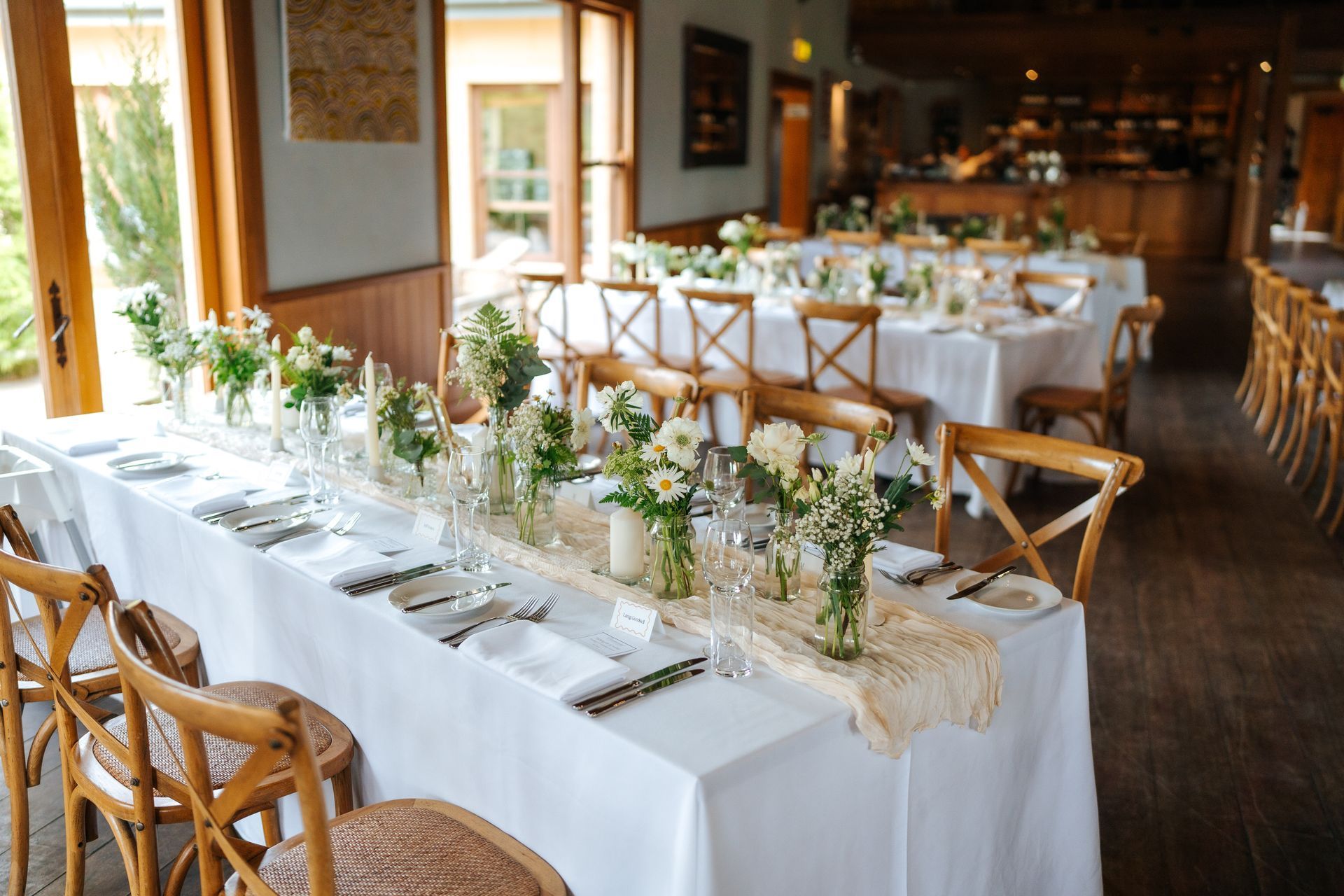 Long table set for a wedding reception with white tablecloth, floral centerpieces, and wooden chairs.