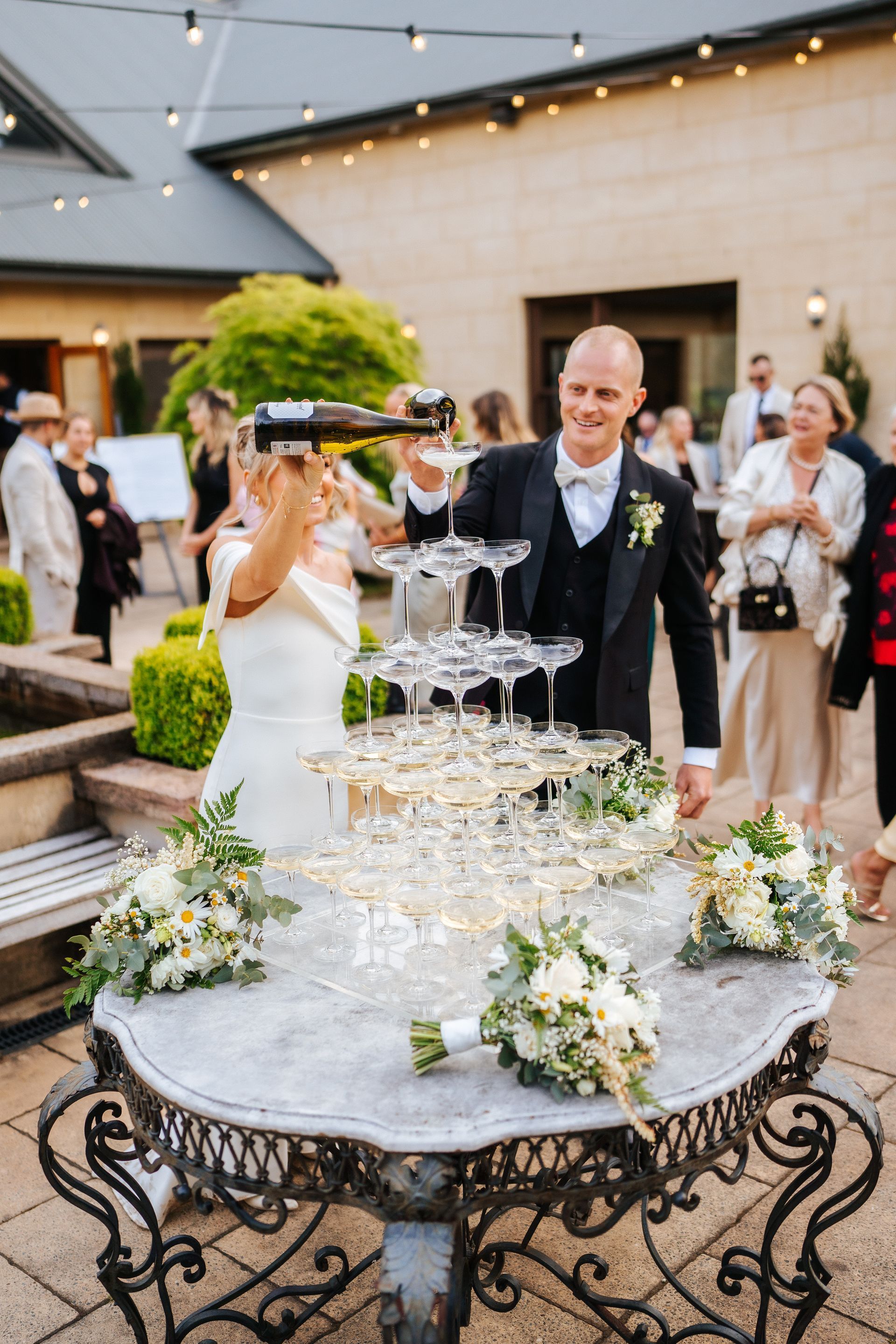 Bride and groom pouring champagne into a tower of glasses at an outdoor wedding reception.