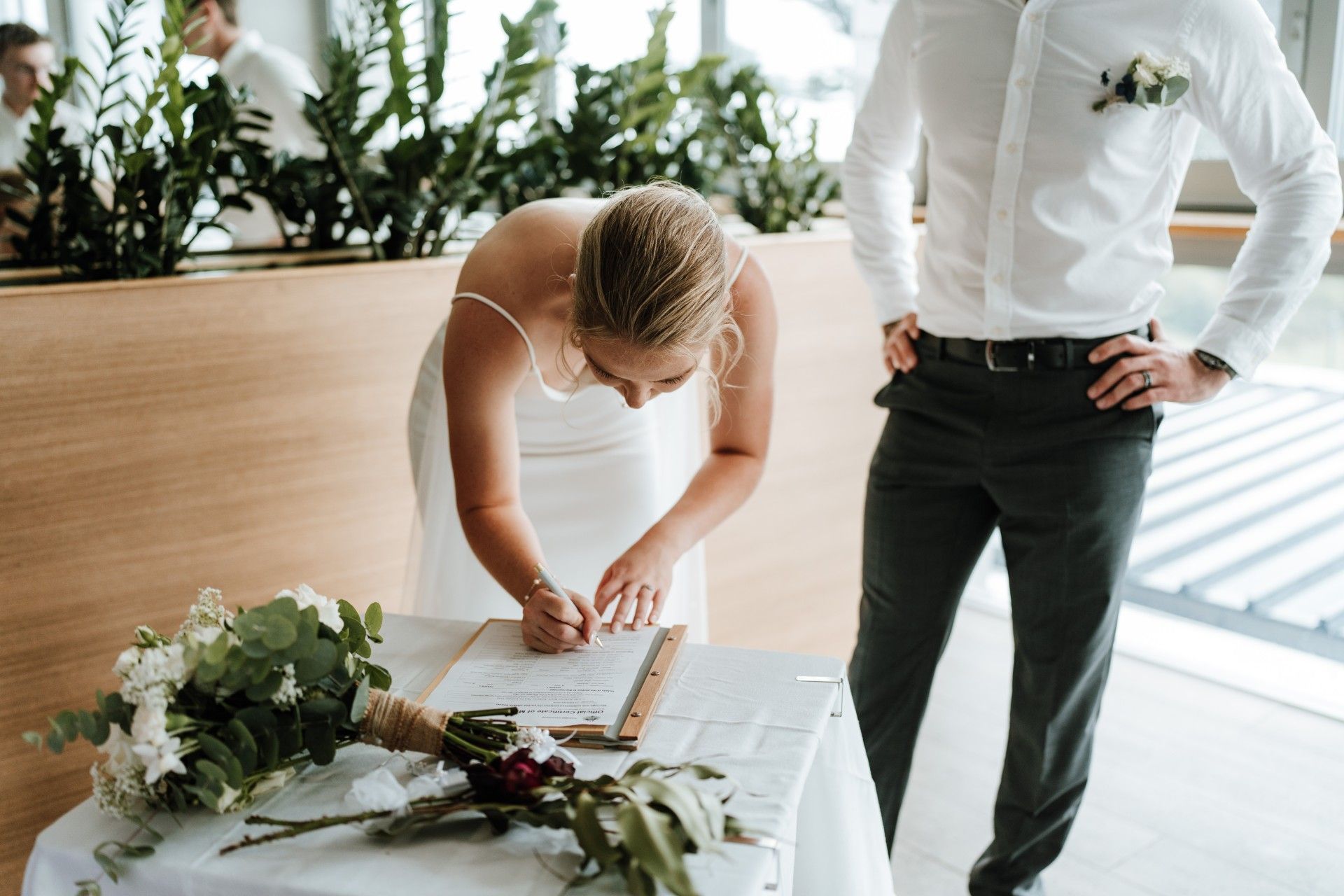 Bride signing marriage certificate at a table, groom watches, flowers in foreground.