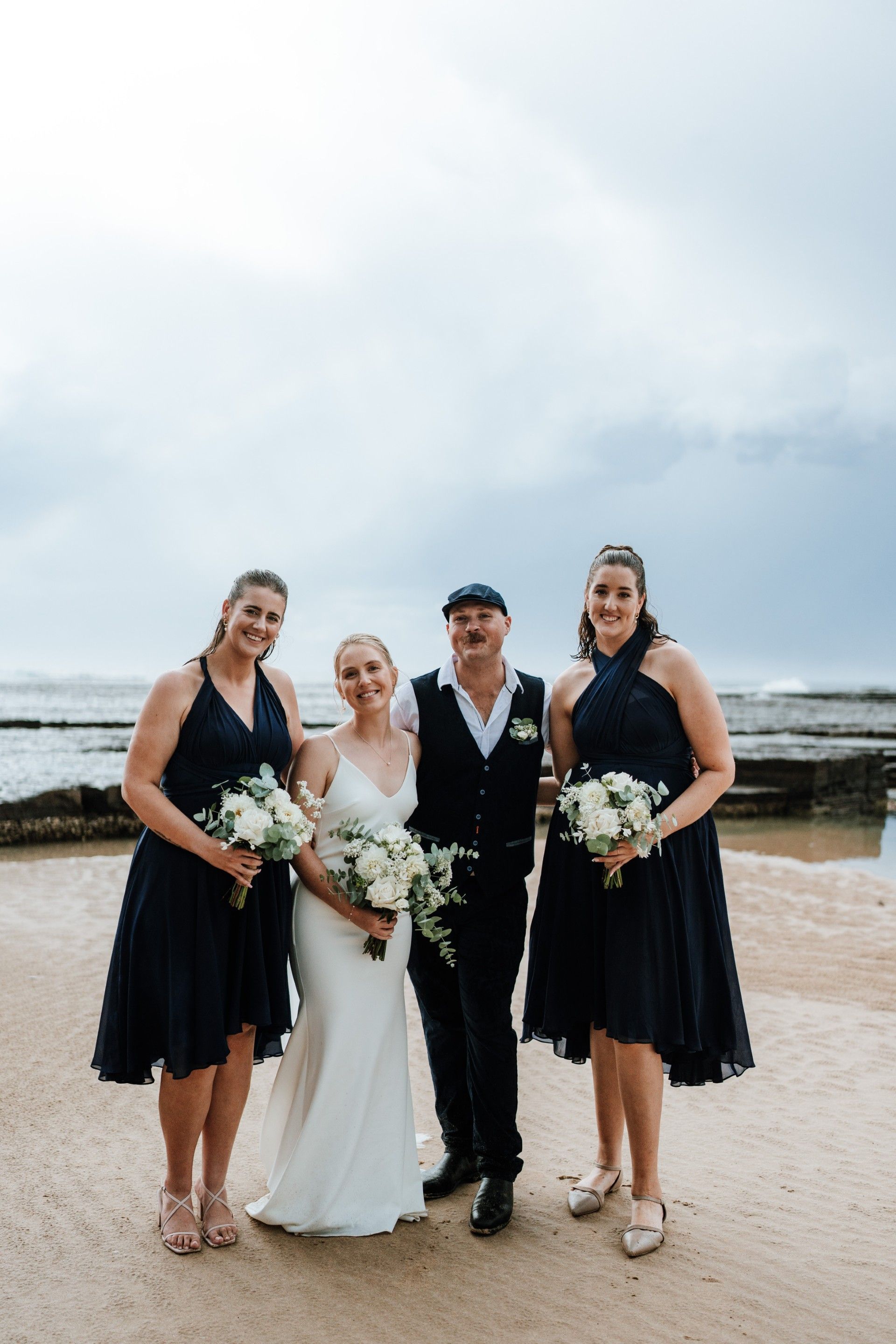Bride and groom with two bridesmaids on a beach, all holding bouquets. Cloudy sky.