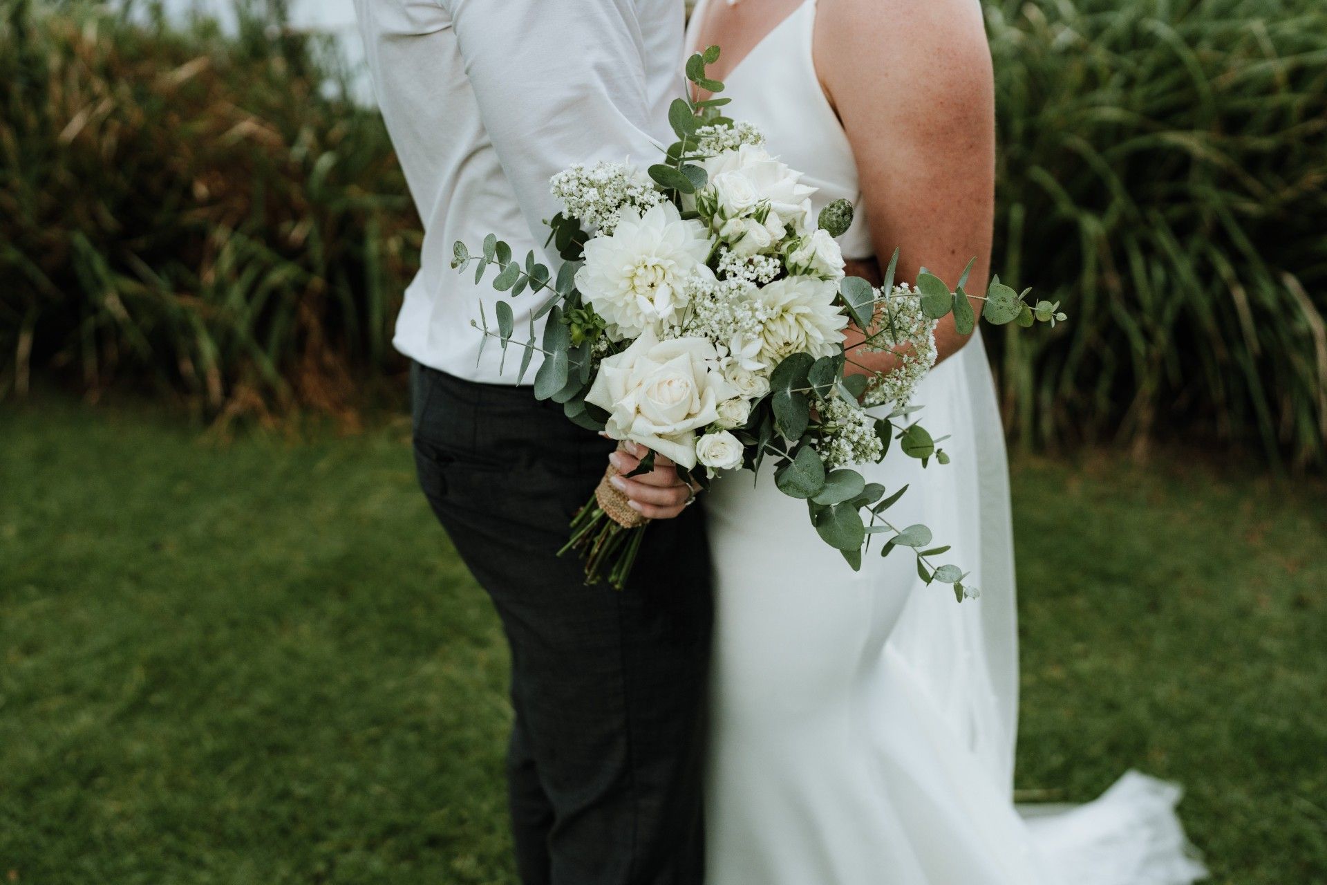Couple embracing, bride holding a white flower bouquet, standing on green grass.