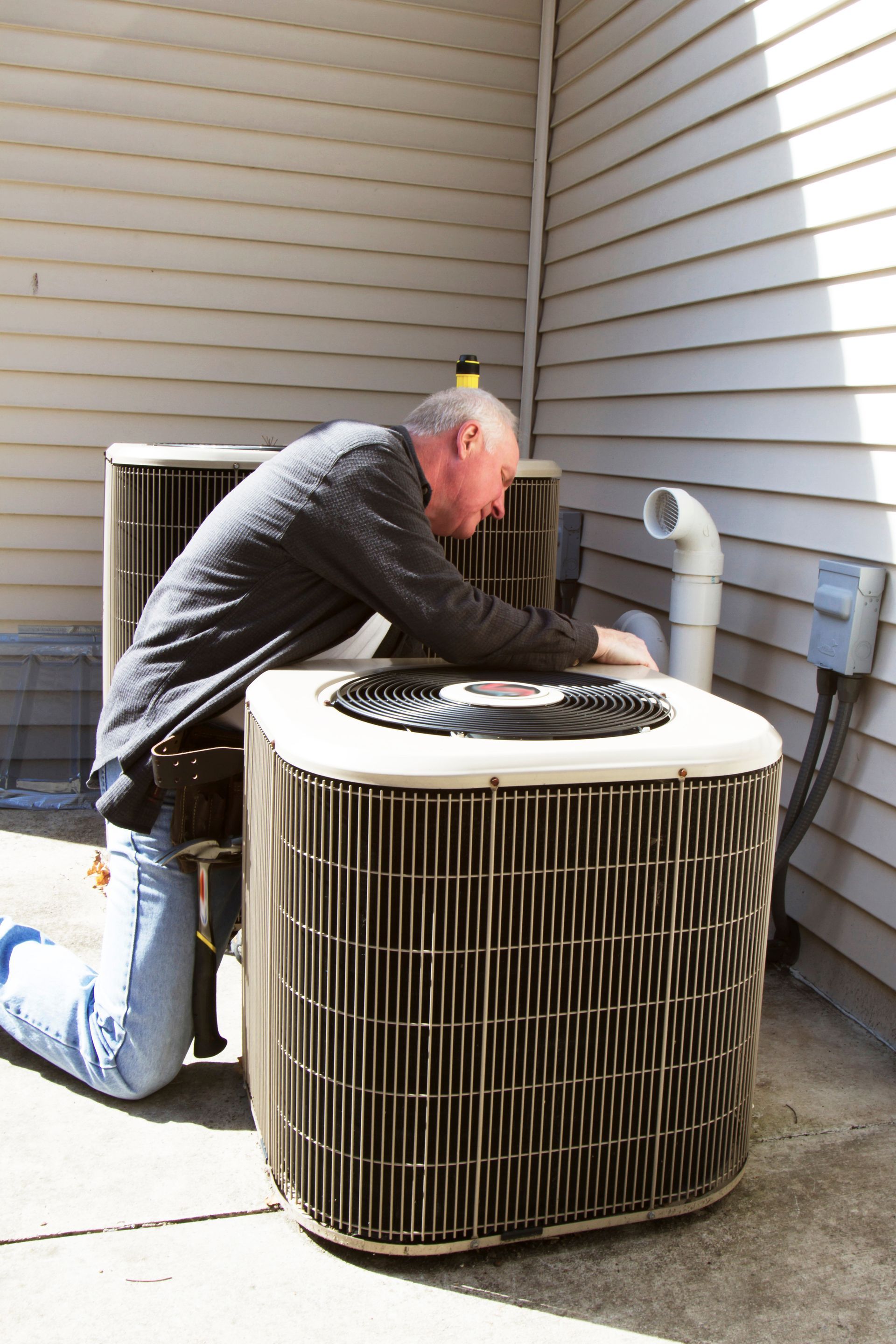 Man kneeling, inspecting an air conditioning unit outside a building.