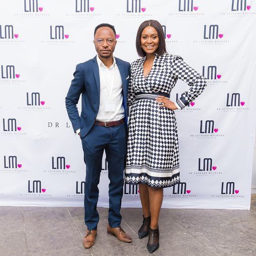 Man and woman posing in front of a banner with the letters LM. The woman wears a checkered dress, and the man wears a suit.
