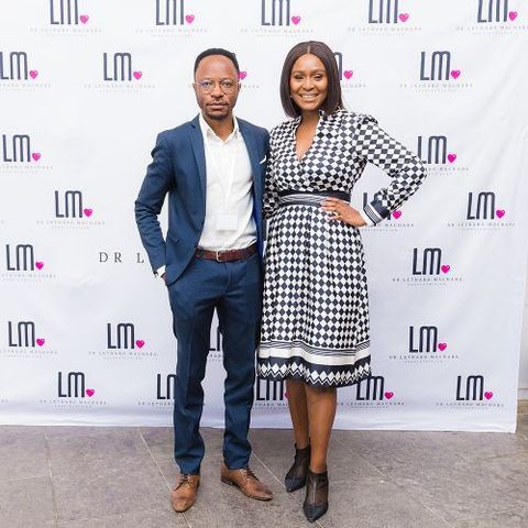Man and woman posing in front of a logo wall. Man in blue suit, woman in black and white dress.