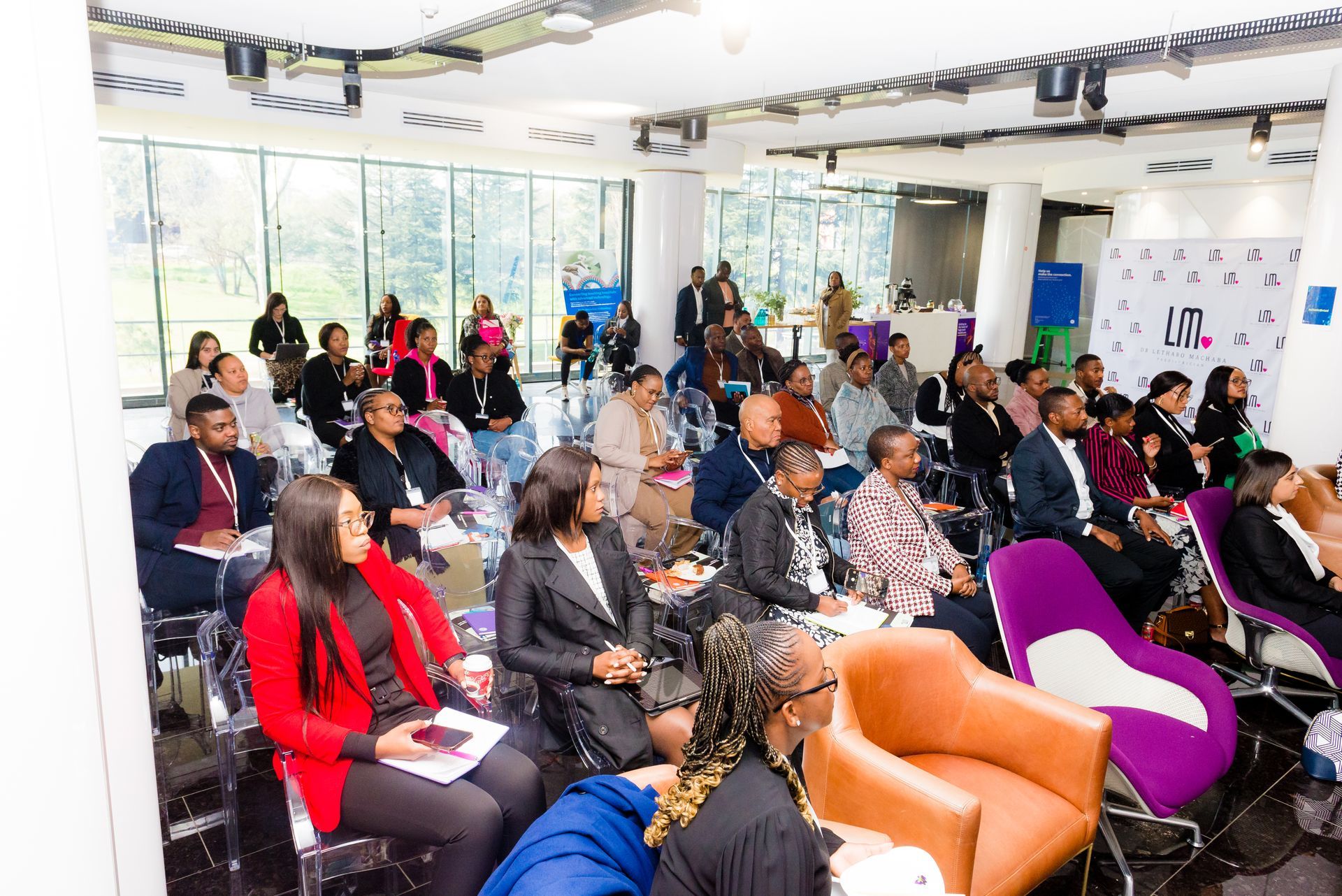 Audience at a conference listening to a presentation in a bright room with large windows.