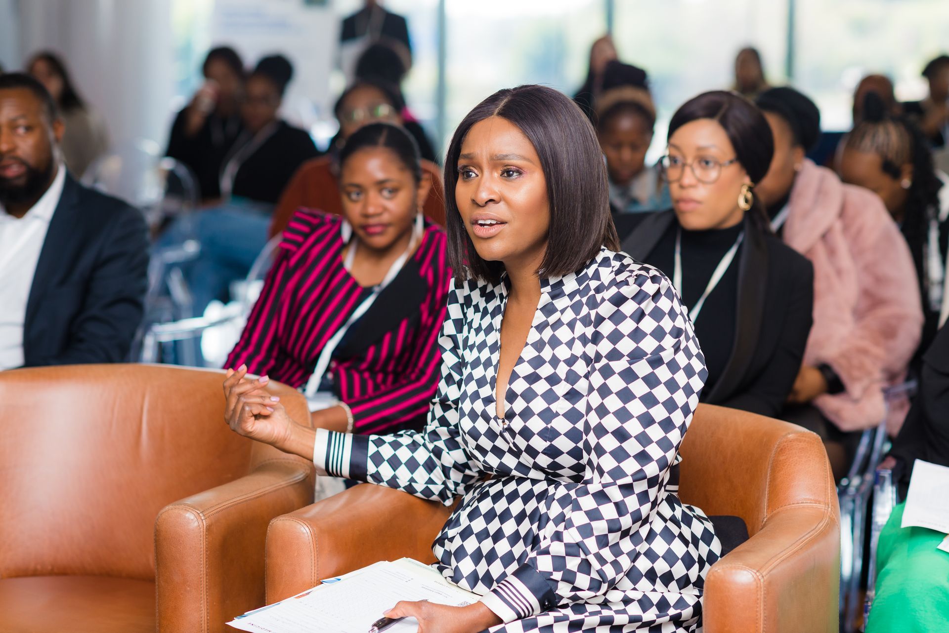 Woman in patterned dress speaking at a conference, other attendees in the background.