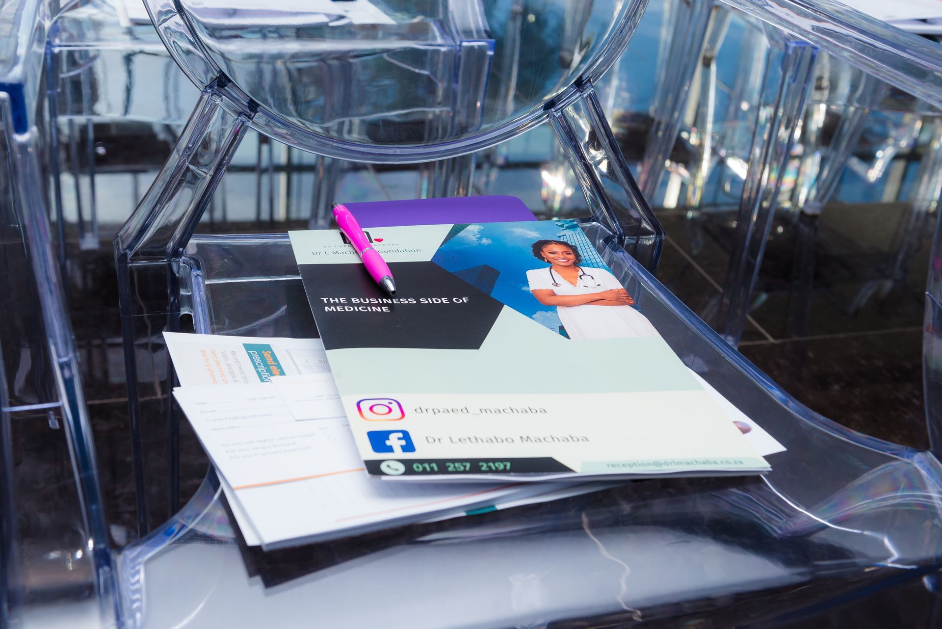 Stack of brochures and business cards in a clear plastic chair; a pink pen rests on top.
