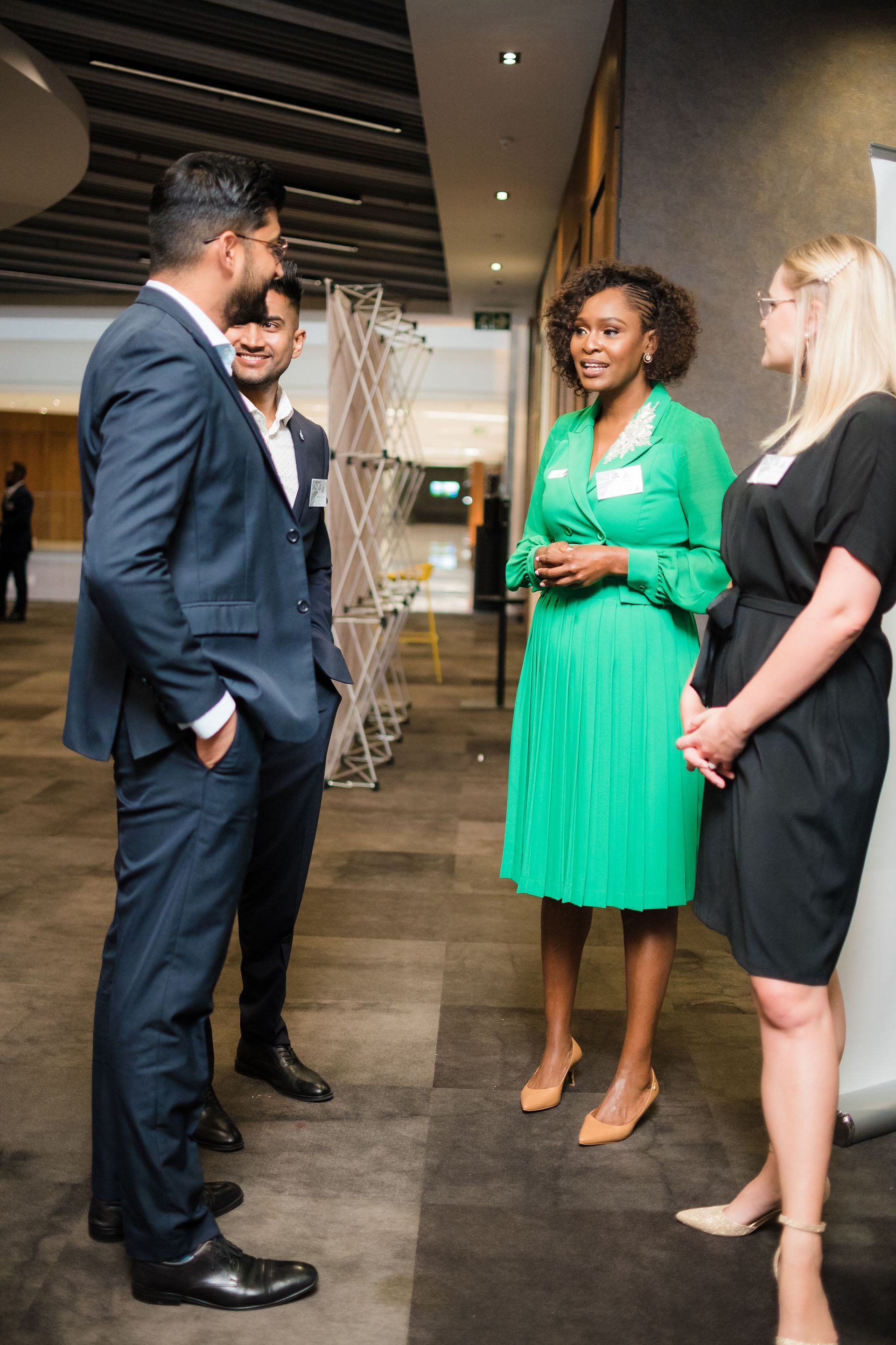 Four people in business attire conversing in an indoor setting. A woman in green dress is speaking.