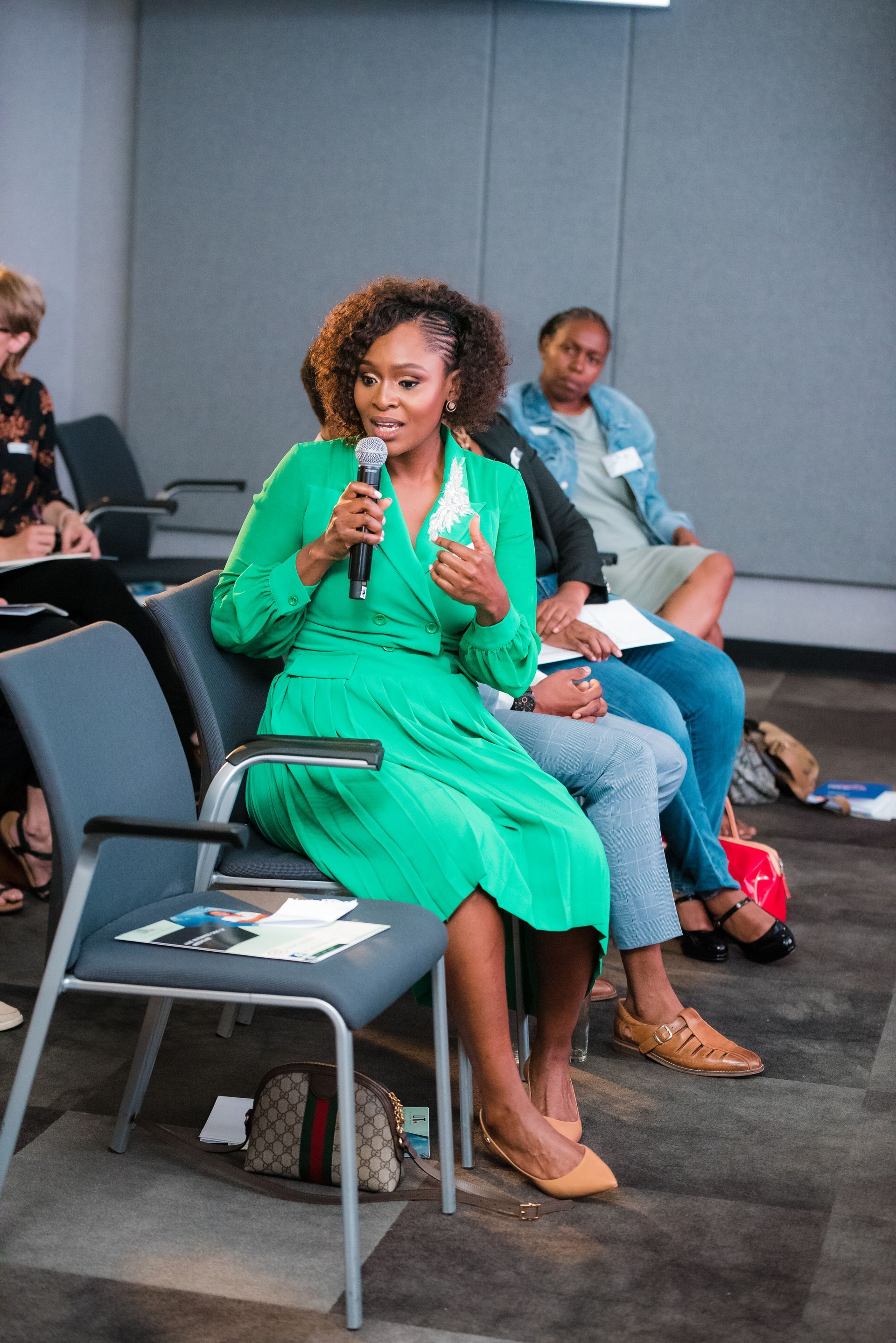 Woman in green dress speaks into a microphone at a conference. Others sit and listen.