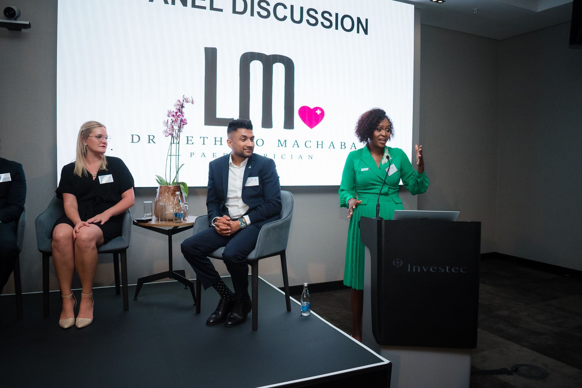 Panel discussion: woman in green speaks at podium, two seated panelists, logo backdrop.