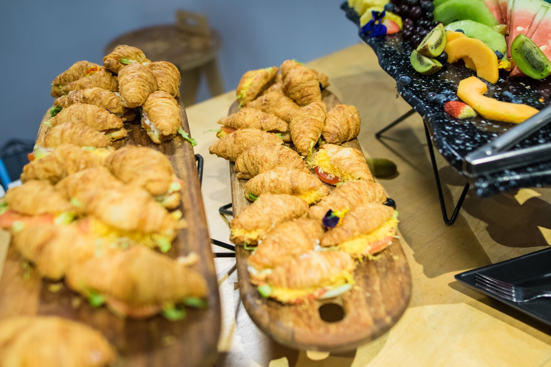 Croissant sandwiches on wooden platters, alongside a fruit platter, at a buffet.