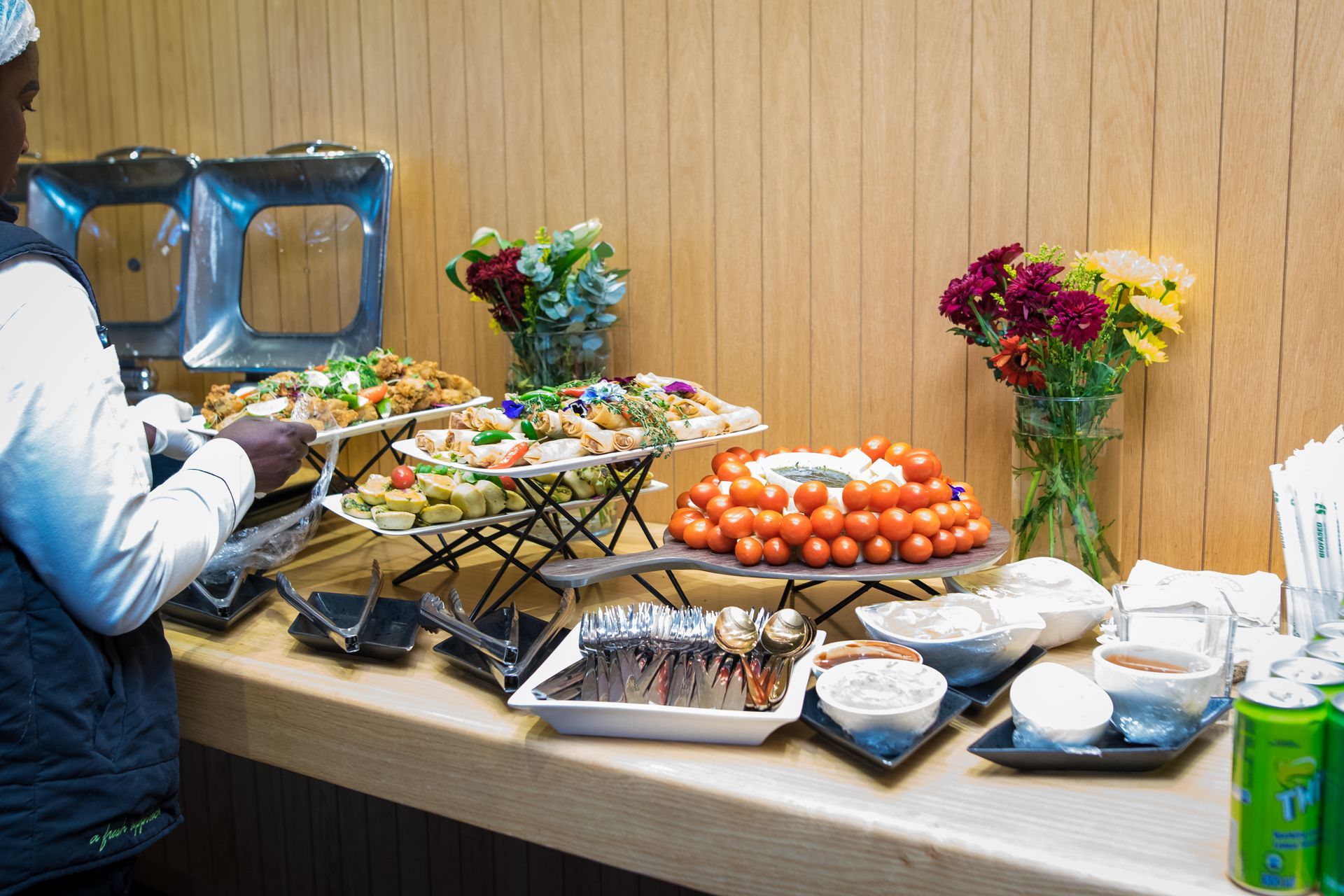 Person at a buffet, selecting food. Tables display various dishes, floral arrangements, and drinks against a wood-paneled wall.