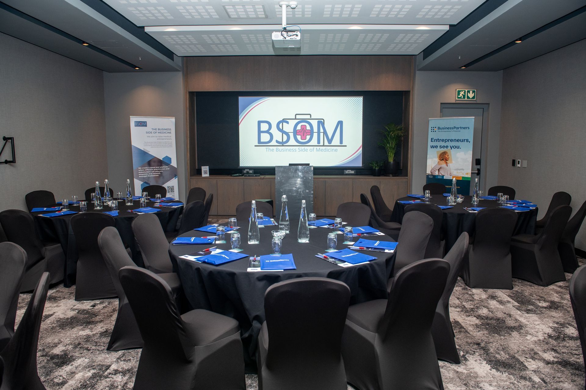 Conference room set for a banquet, with round tables, black chairs, blue table settings, and a large presentation screen.