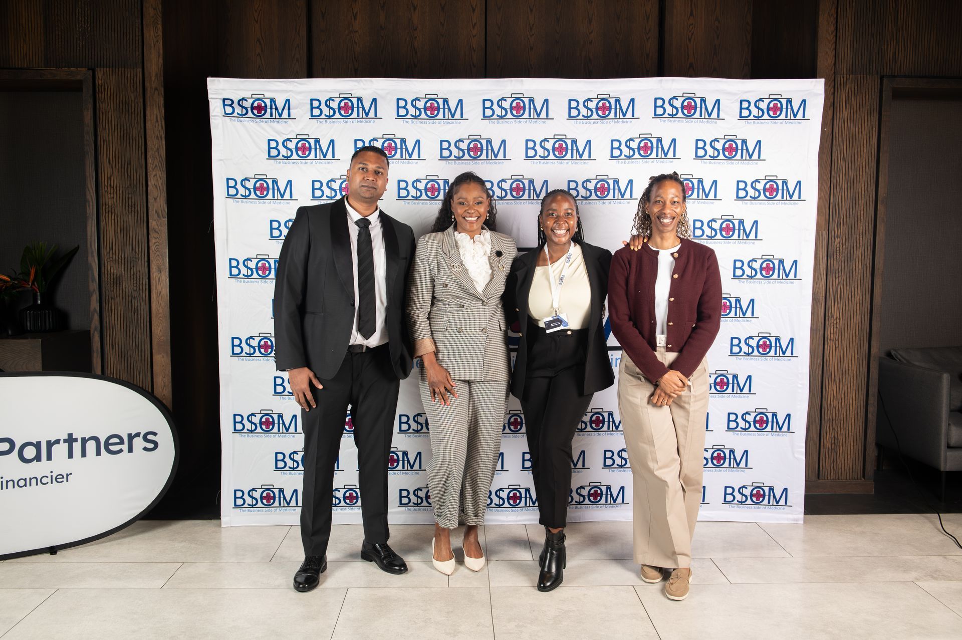 Four people posing in business attire at a branded event backdrop with a “Partners” sign nearby.