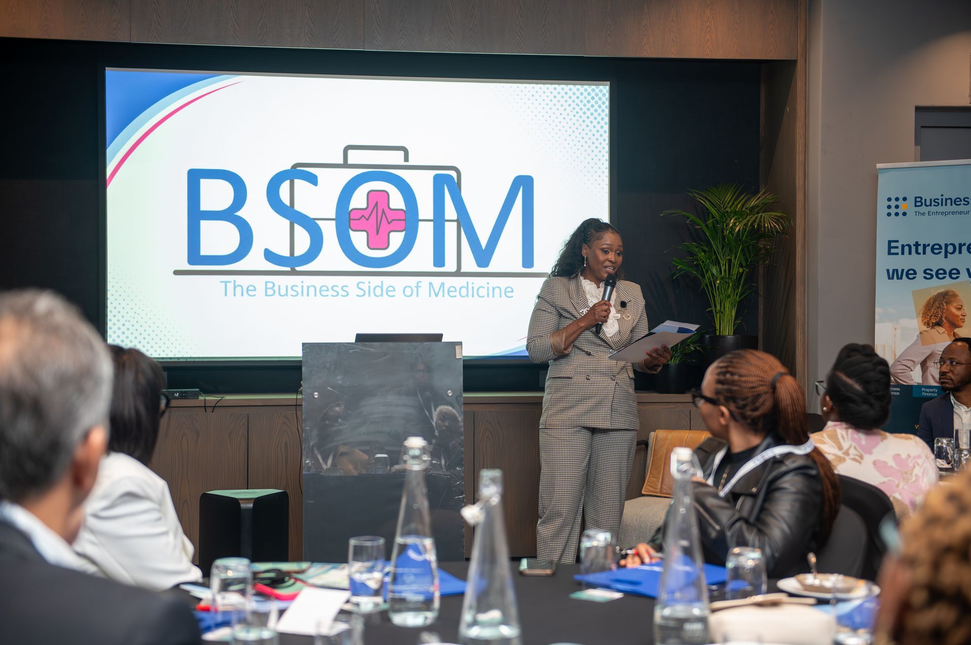 Presenter speaking at a conference beside a BSOM slide on a large screen