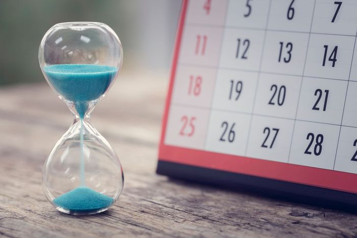An hourglass is sitting on a wooden table next to a calendar.