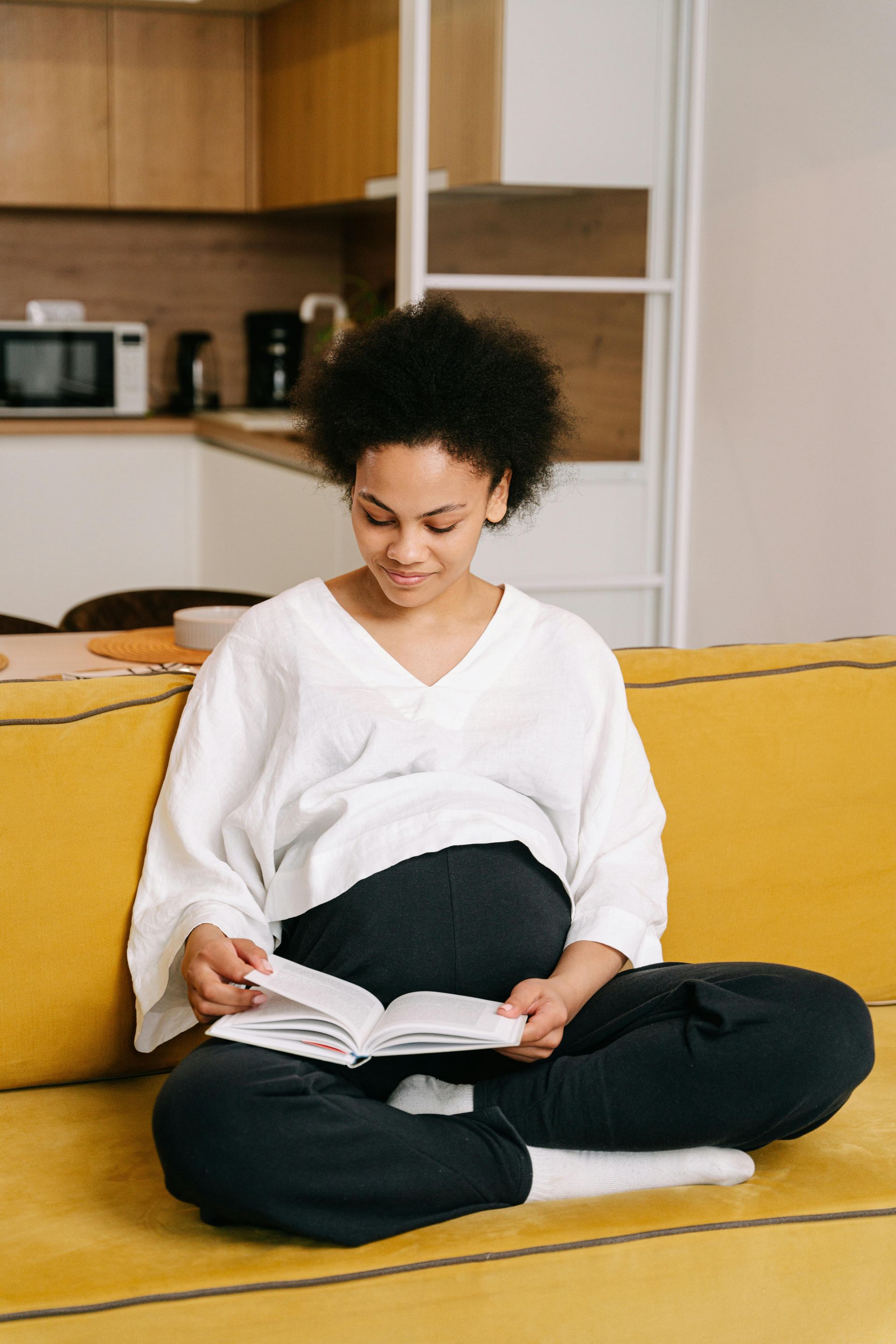 A pregnant woman is sitting on a yellow couch reading a book.
