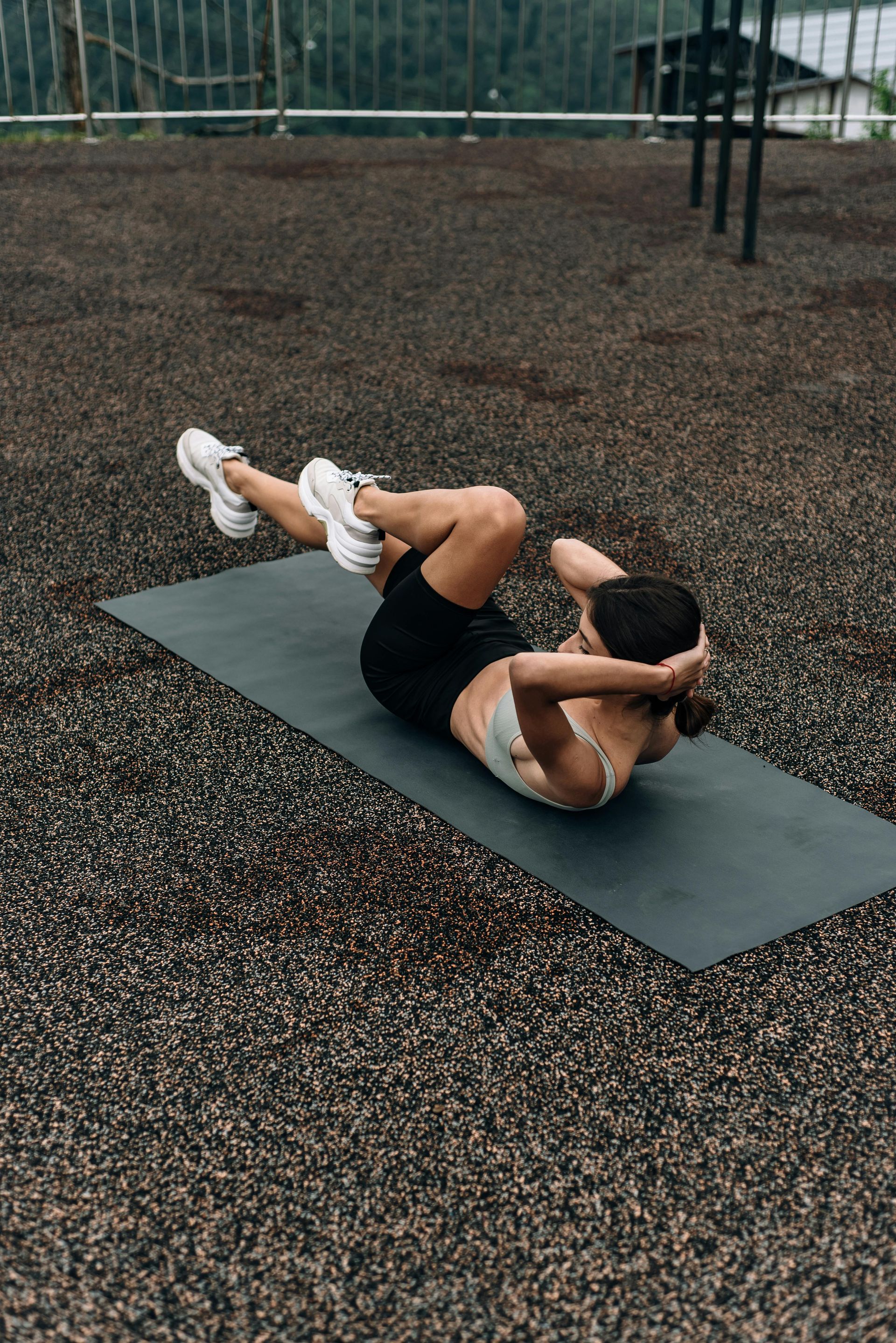 Woman doing bicycle crunches on a mat outdoors. Wearing a sports bra, shorts and sneakers.