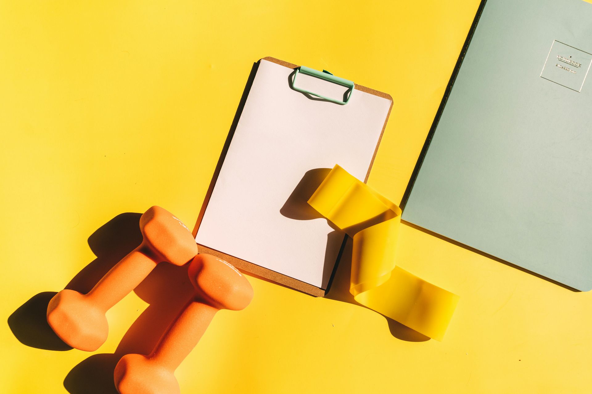 A clipboard , dumbbells , and a notebook are on a yellow table.