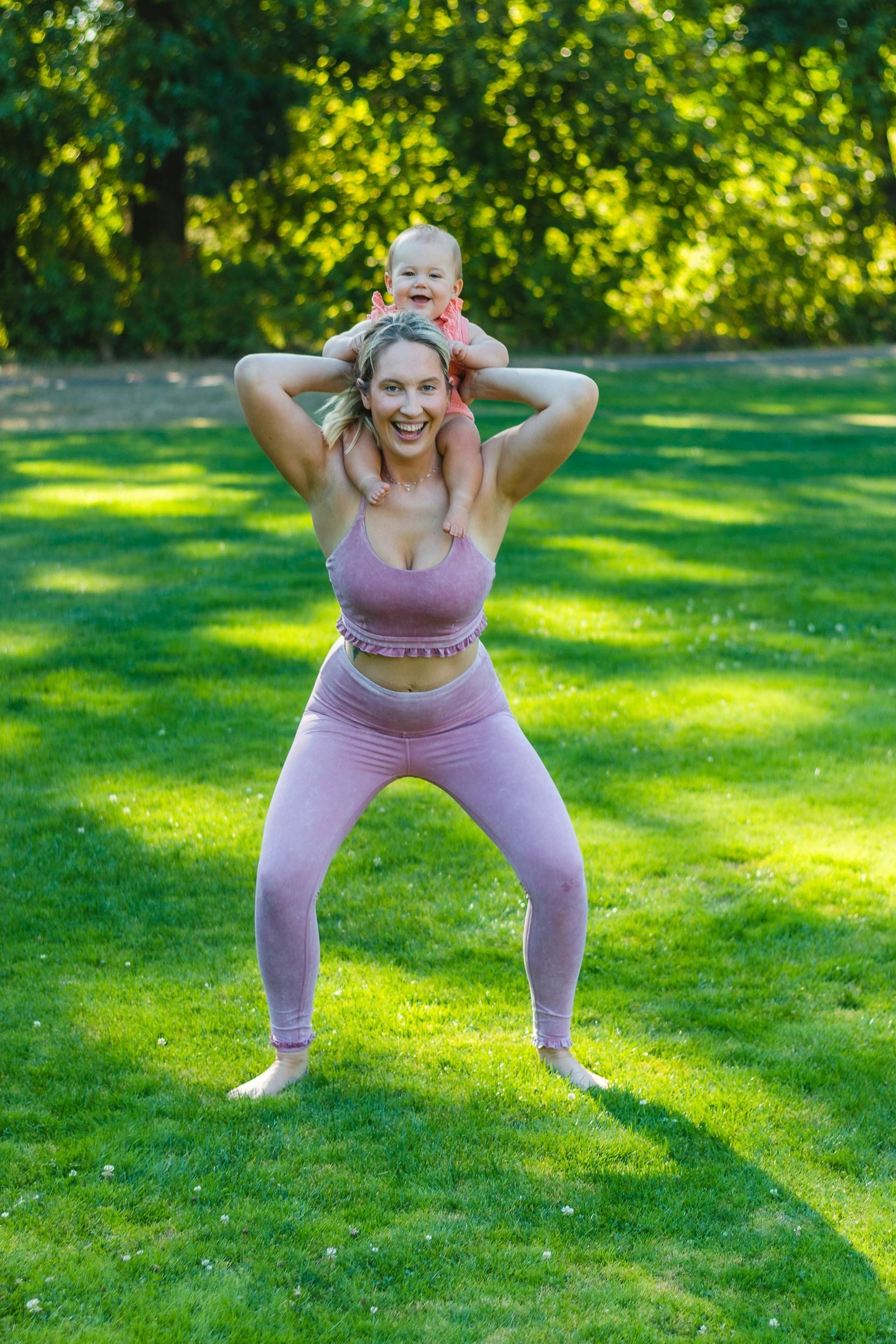 Woman squatting with baby on shoulders in a park, both smiling, wearing pink activewear.
