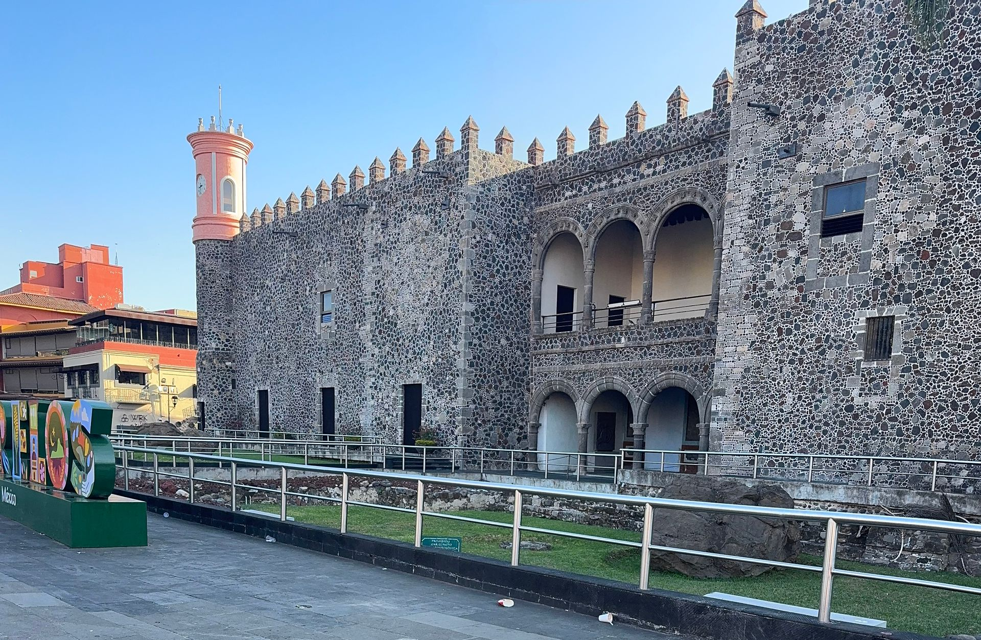 Stone fortress with arched openings, a pink tower, and a guardrail, set against a blue sky.