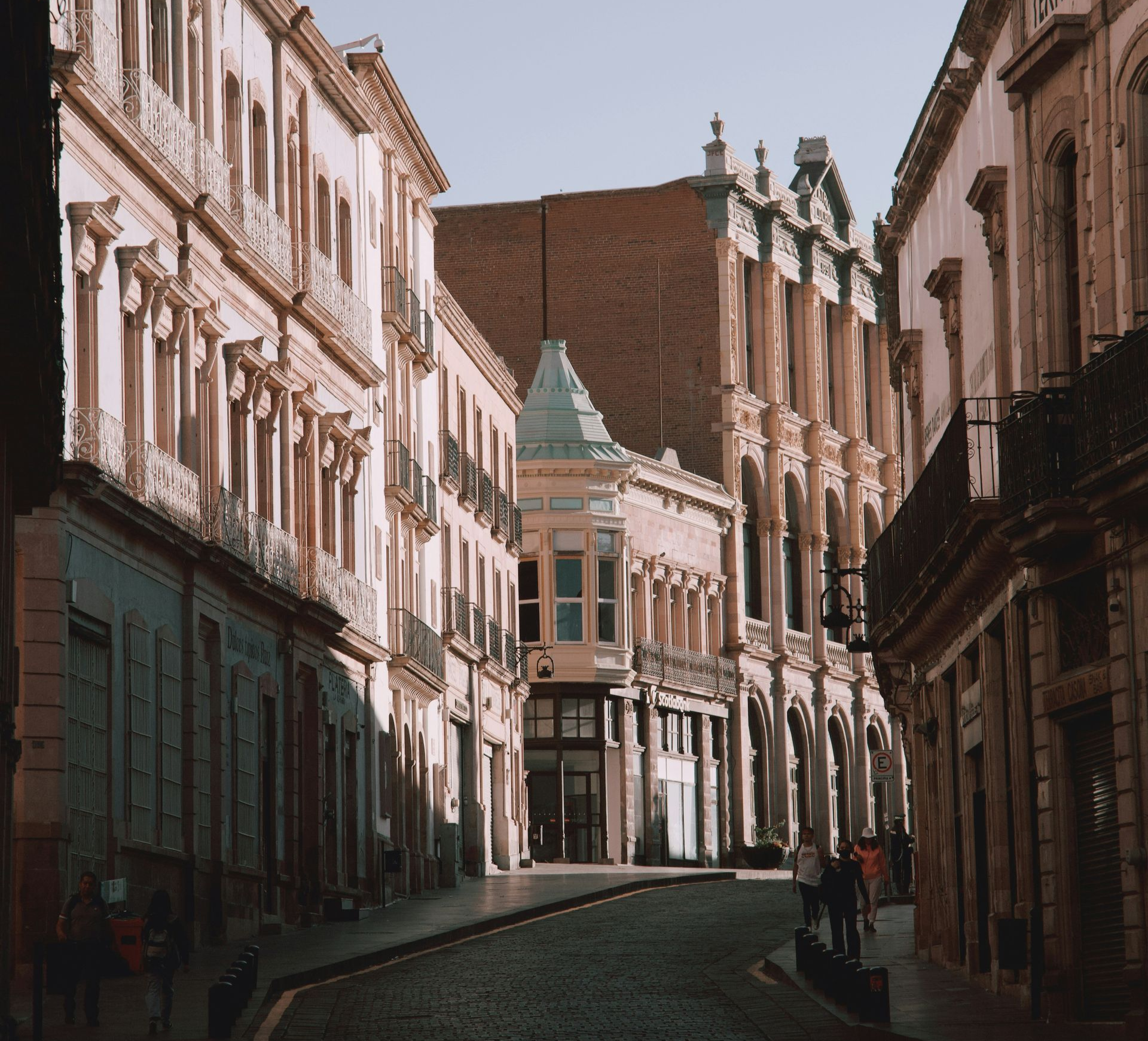 Cobblestone street lined with light-colored buildings; a few people walk in the distance.