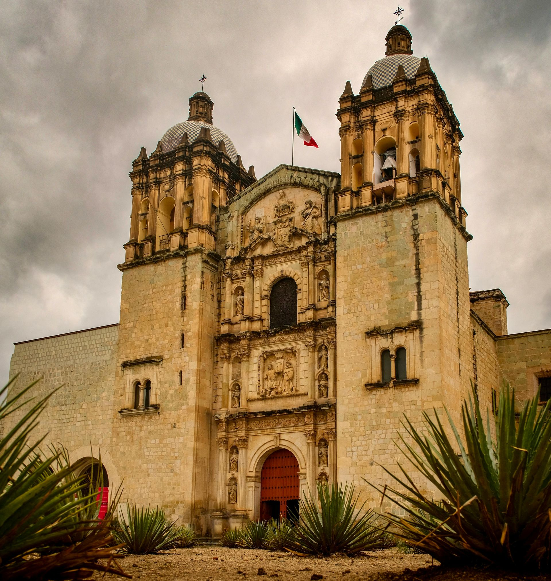 Stone cathedral with two bell towers, red door, Mexican flag, cloudy sky.