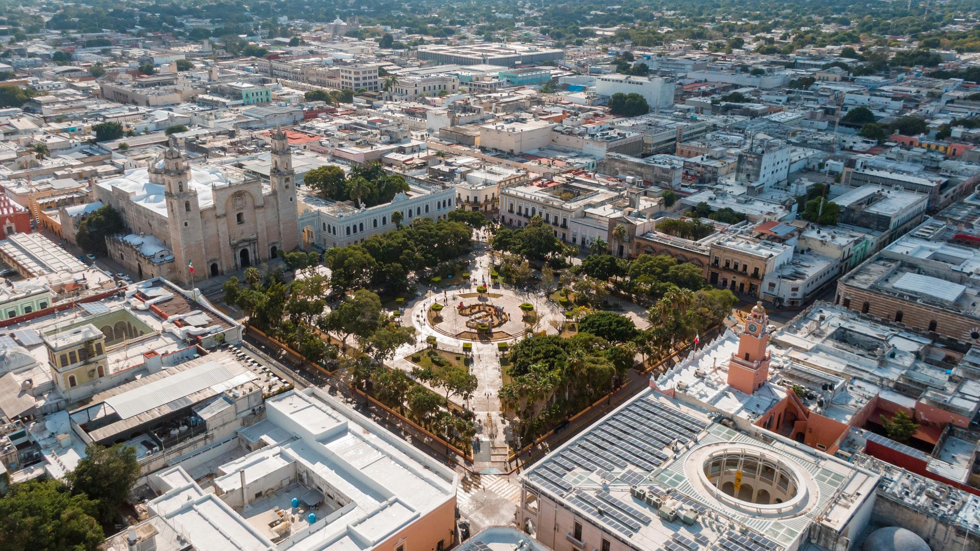 Aerial view of Mérida, Mexico's city center, featuring a large plaza with trees, surrounded by buildings and a cathedral.